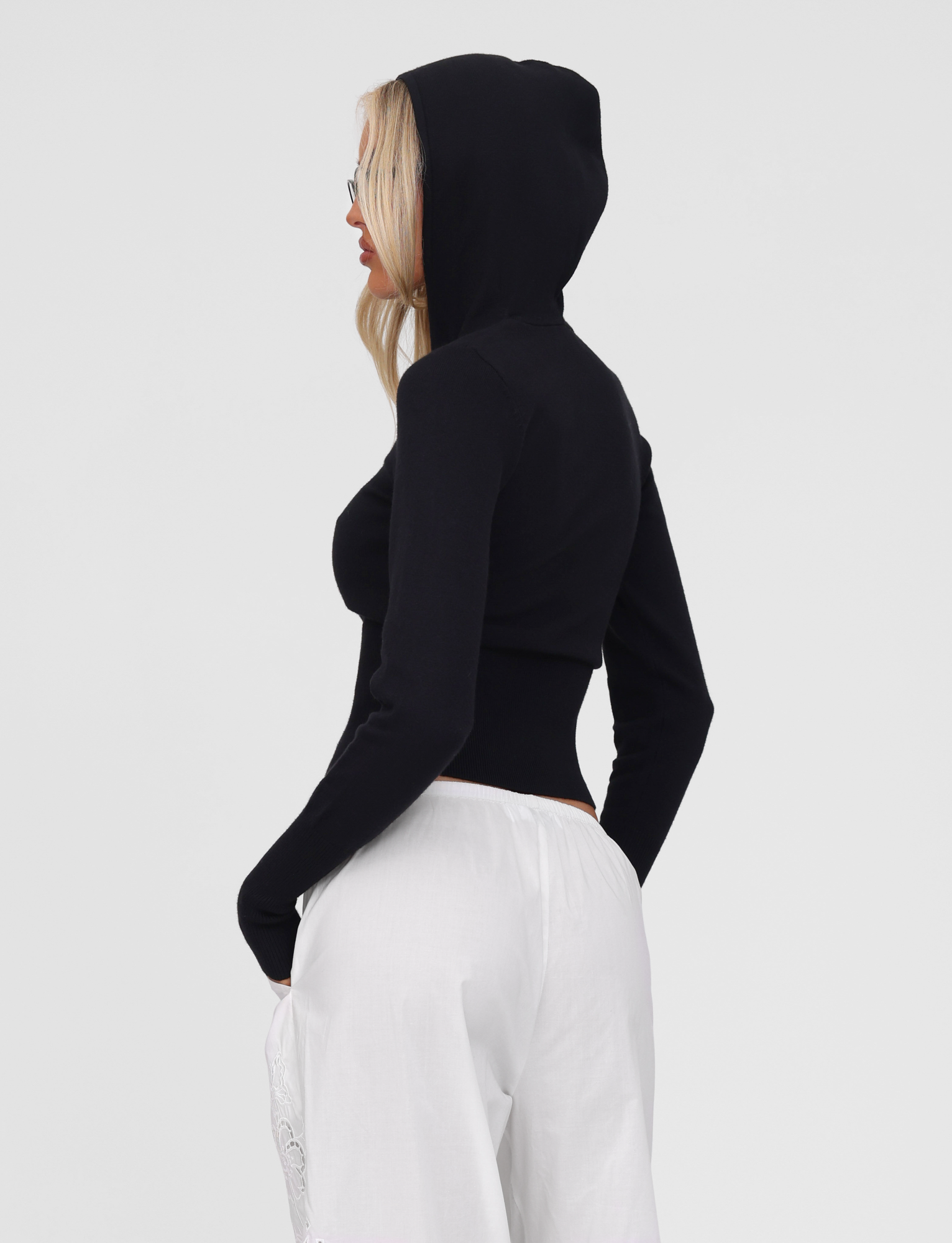 Wearing the TIGERMIST SARINA JUMPER in black, a person stands in profile against a white background, hands in pockets, with blonde hair peeking from under the hood and paired with loose white pants.