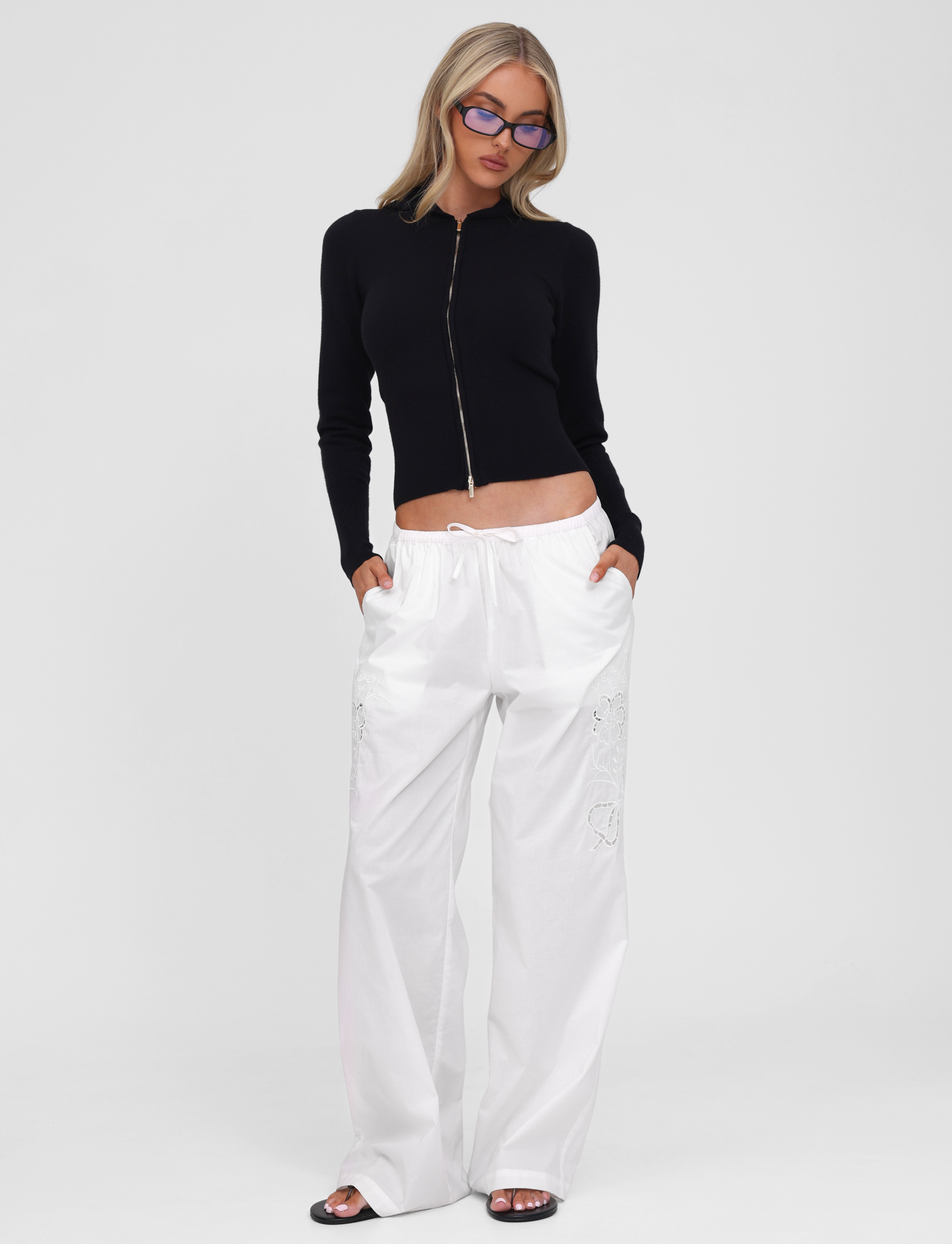 A person stands against a plain white background with hands in pockets, wearing the SARINA JUMPER - BLACK by TIGERMIST, paired with loose white drawstring pants, black sandals, and blue-tinted glasses.