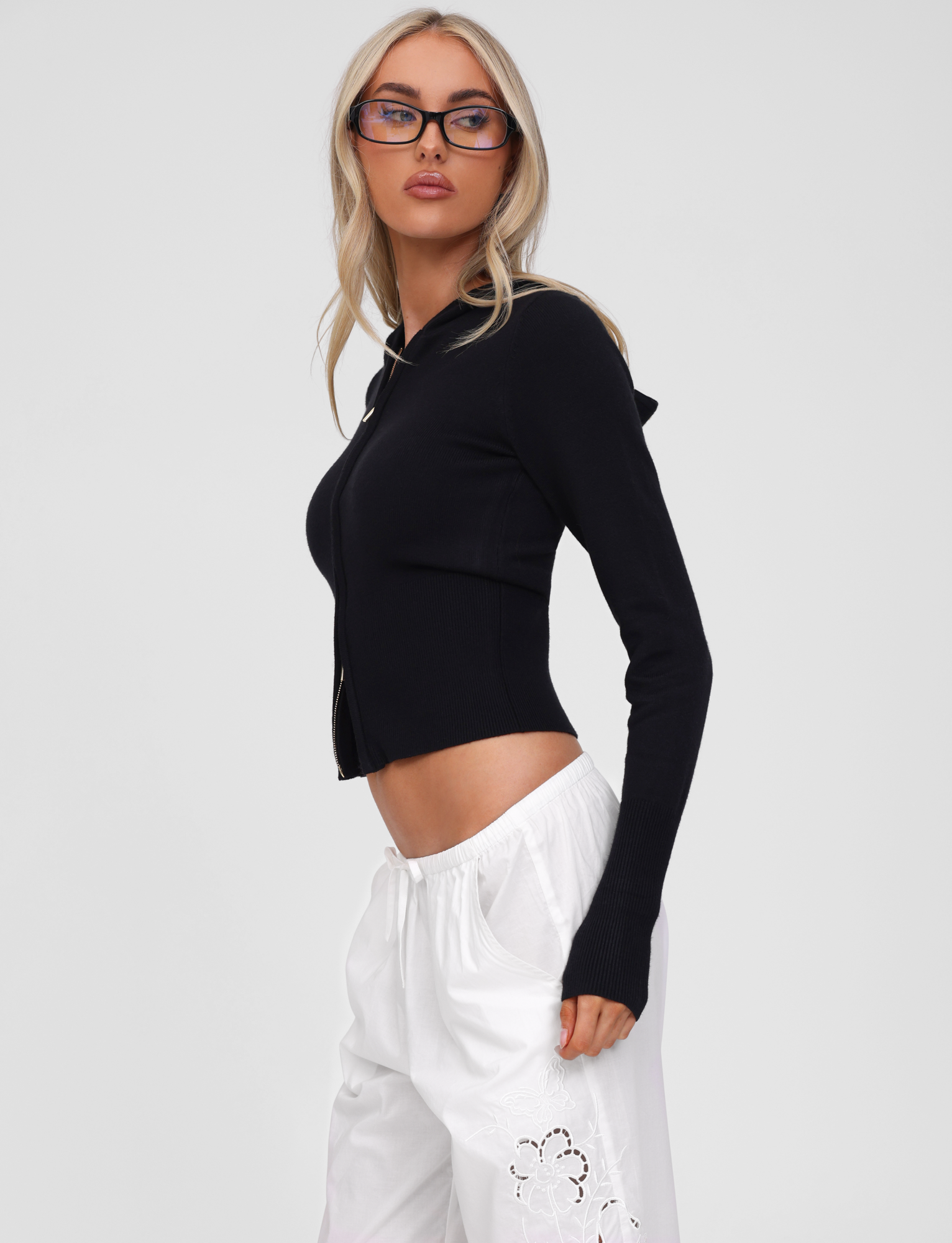 A woman with long blonde hair and glasses models the TIGERMIST SARINA JUMPER in black—a fitted knit hoodie with a gold zipper—paired with loose white pants featuring floral cutouts, set against a plain light gray background.