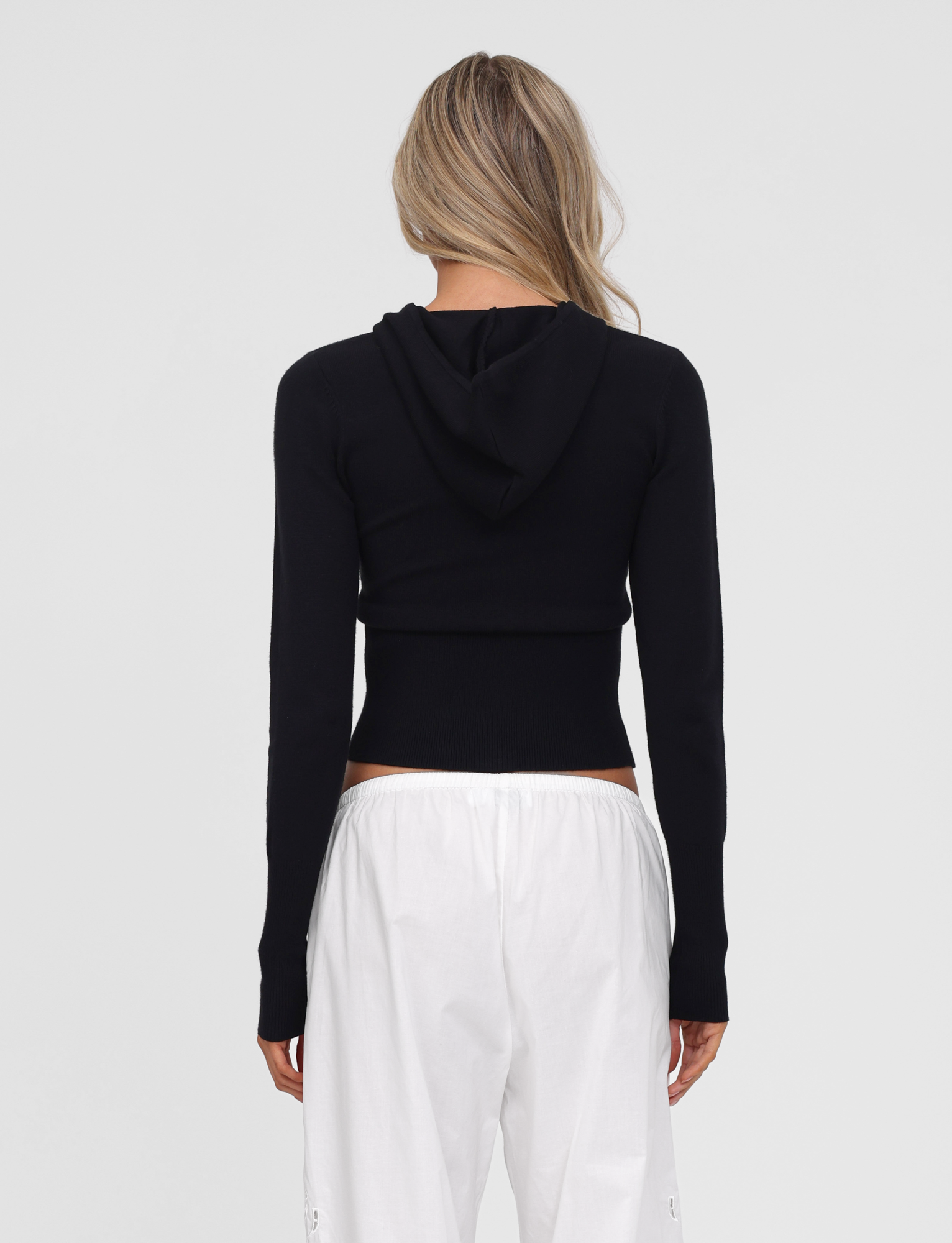 A person with long, wavy blonde hair stands with their back to the camera, wearing the TIGERMIST SARINA JUMPER in black and loose white pants against a plain light background.