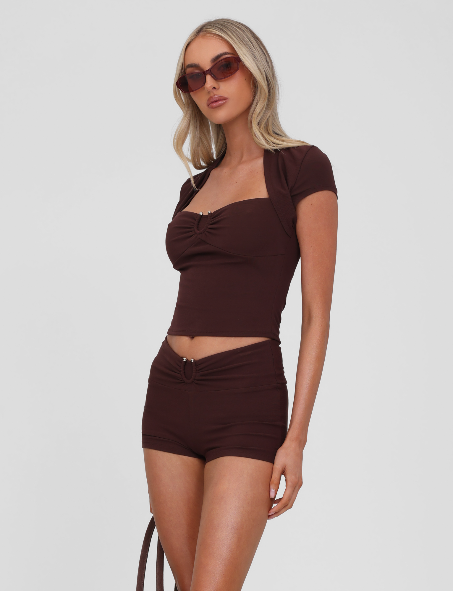 A woman wears the TIGERMIST LUNARA TOP in chocolate with matching high-waisted shorts and brown sunglasses, standing against a plain white background.
