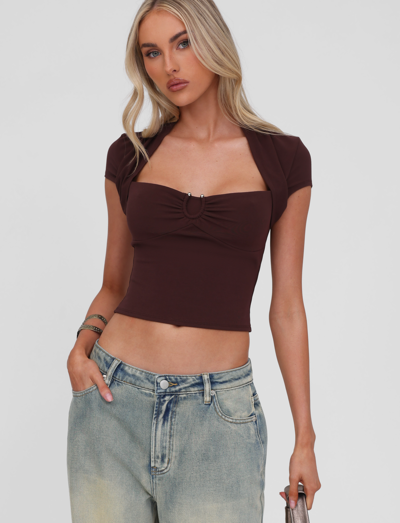 A woman with long blonde hair wears the TIGERMIST LUNARA TOP in chocolate, a fitted short-sleeve style, paired with light-wash high-waisted jeans. She holds a phone and stands against a plain background, her expression neutral.