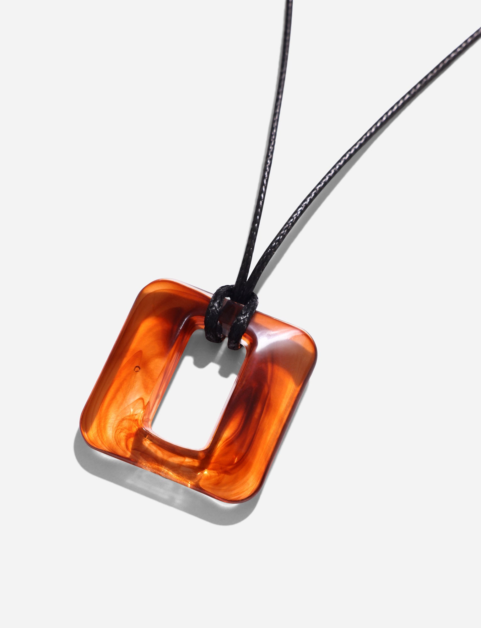 The TIGERMIST MIRREN SQUARE NECKLACE - BROWN features a minimalist square-shaped translucent amber pendant with rounded edges, a central cutout, and a glossy finish. It hangs on a thin woven cord elegantly tied through the top, set against a plain white background.