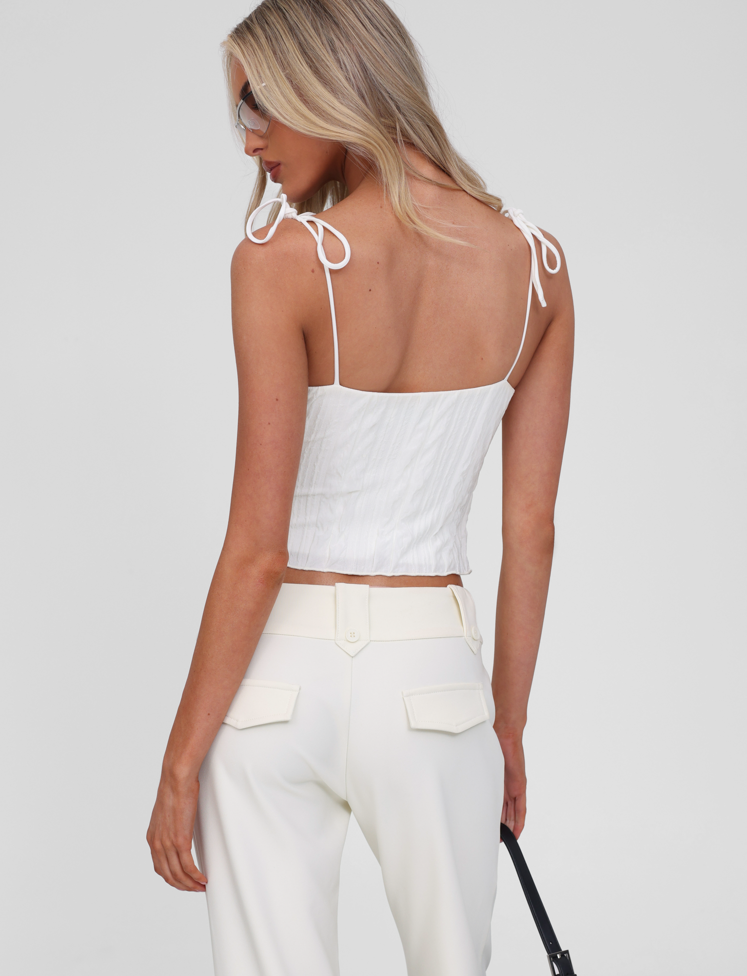 A woman with long blonde hair wears the TIGERMIST RANIA TOP in off-white and white pants, facing away while looking over her shoulder and holding a black strap.