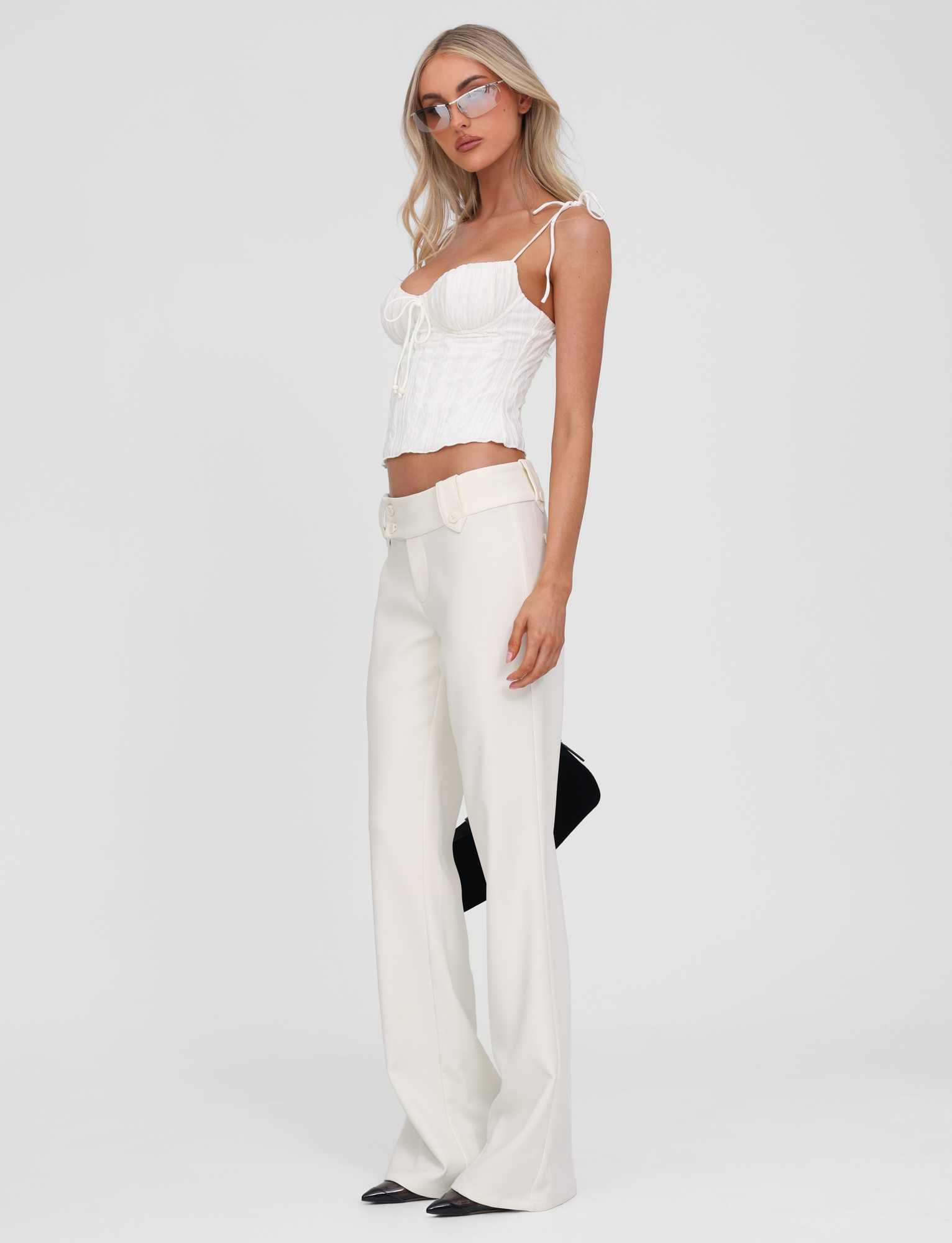 A woman with long blonde hair and glasses stands against a white background wearing the TIGERMIST RANIA TOP in off-white, featuring strappy shoulders and a sweetheart neckline, paired with high-waisted wide-leg white pants and holding a black clutch.