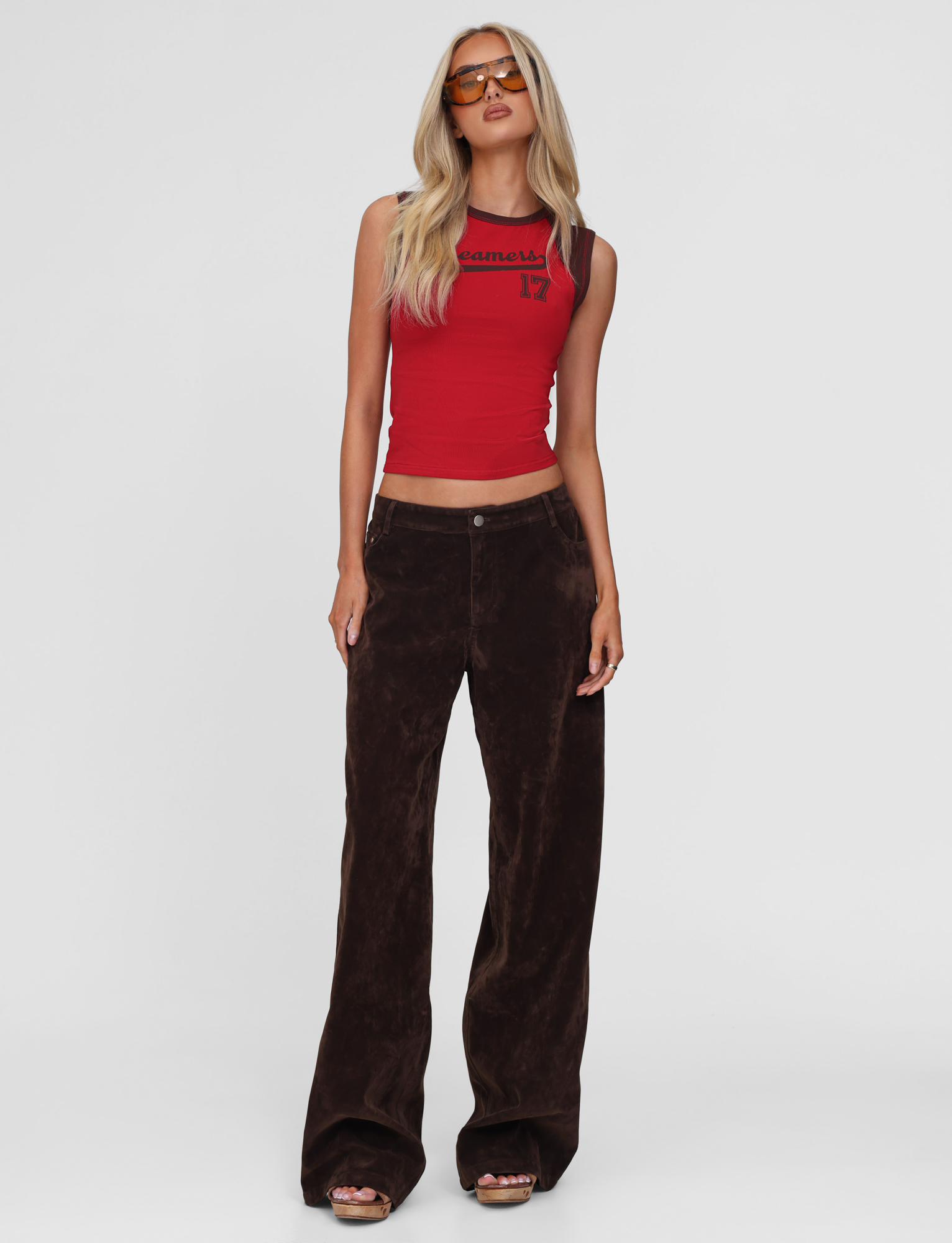 A woman wearing the TIGERMIST FIERA TEE - RED crop top, loose dark brown pants, brown sandals, and amber-tinted sunglasses stands against a plain white background.