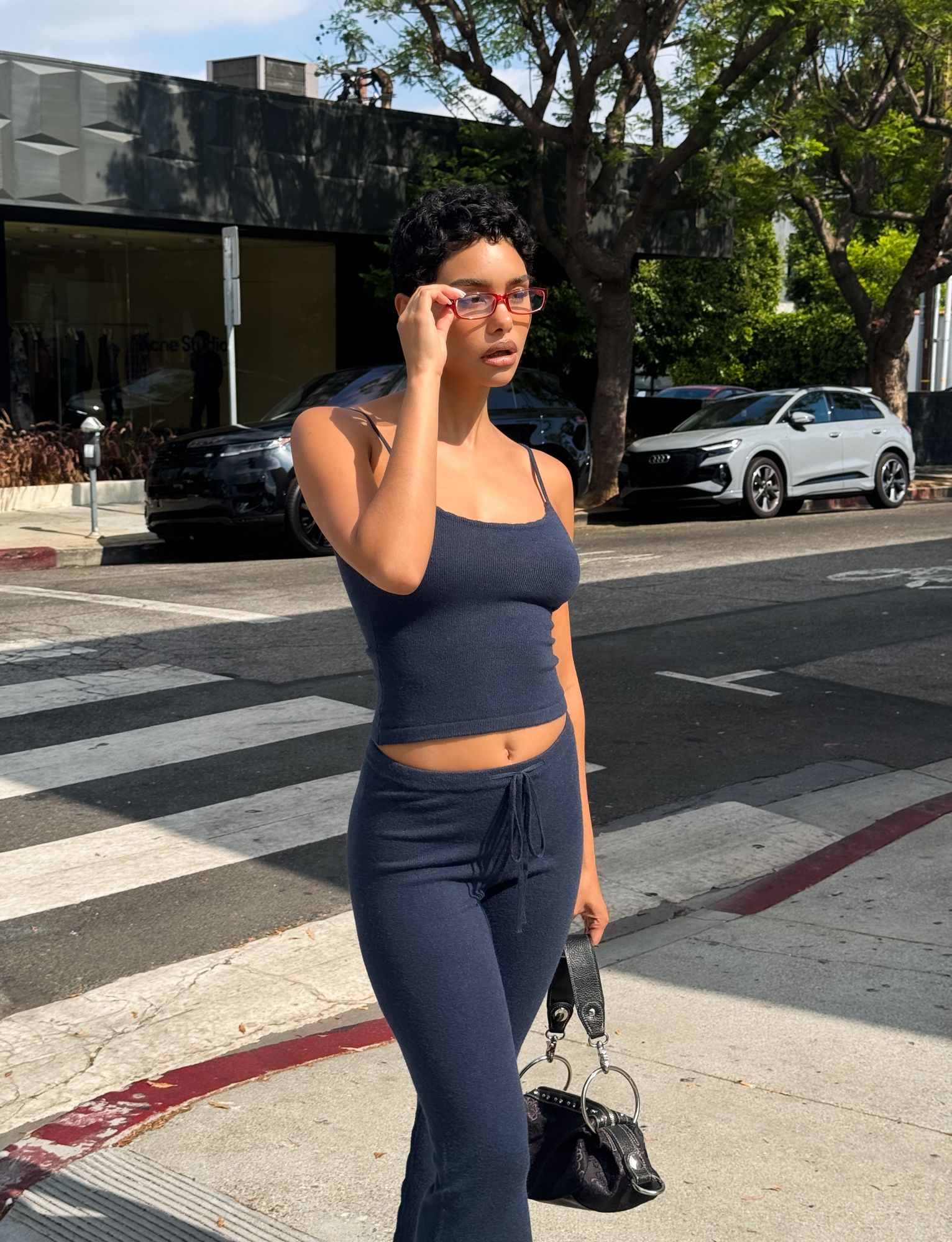 Wearing the TIGERMIST LEORA TOP - NAVY with adjustable straps and matching pants, a person stands on a city sidewalk holding a black handbag and adjusting red sunglasses. Cars, trees, and a crosswalk are in the background.