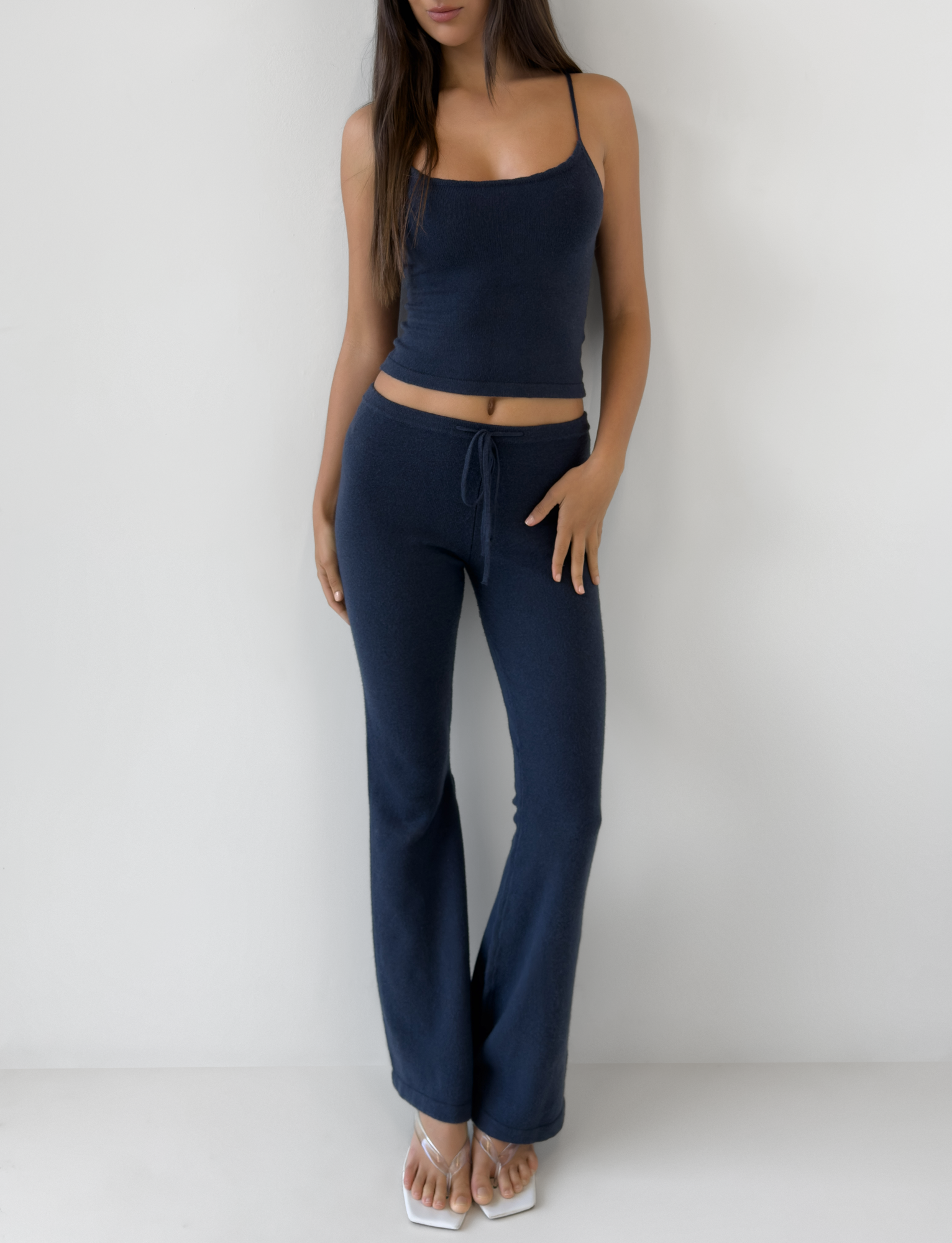 A woman with long hair stands barefoot against a white wall, wearing TIGERMIST's LEORA PANT - NAVY with a drawstring waist and a navy sleeveless crop top for an effortless look, her right hand resting on her thigh.