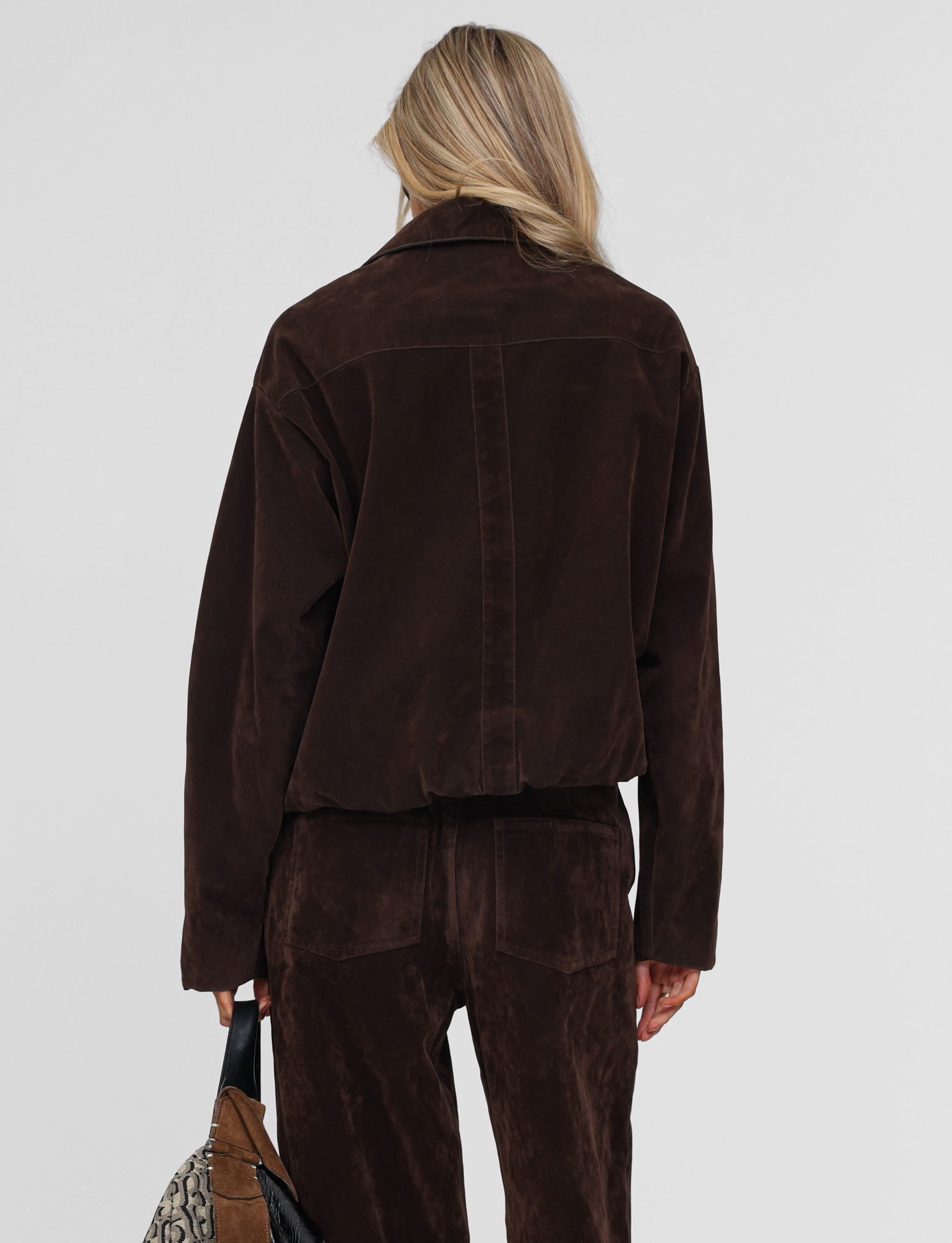 A person with long blonde hair is seen from behind wearing the TIGERMIST CAMELLIA BOMBER JACKET in chocolate suede, paired with matching pants and holding a snakeskin-patterned bag against a plain light background.