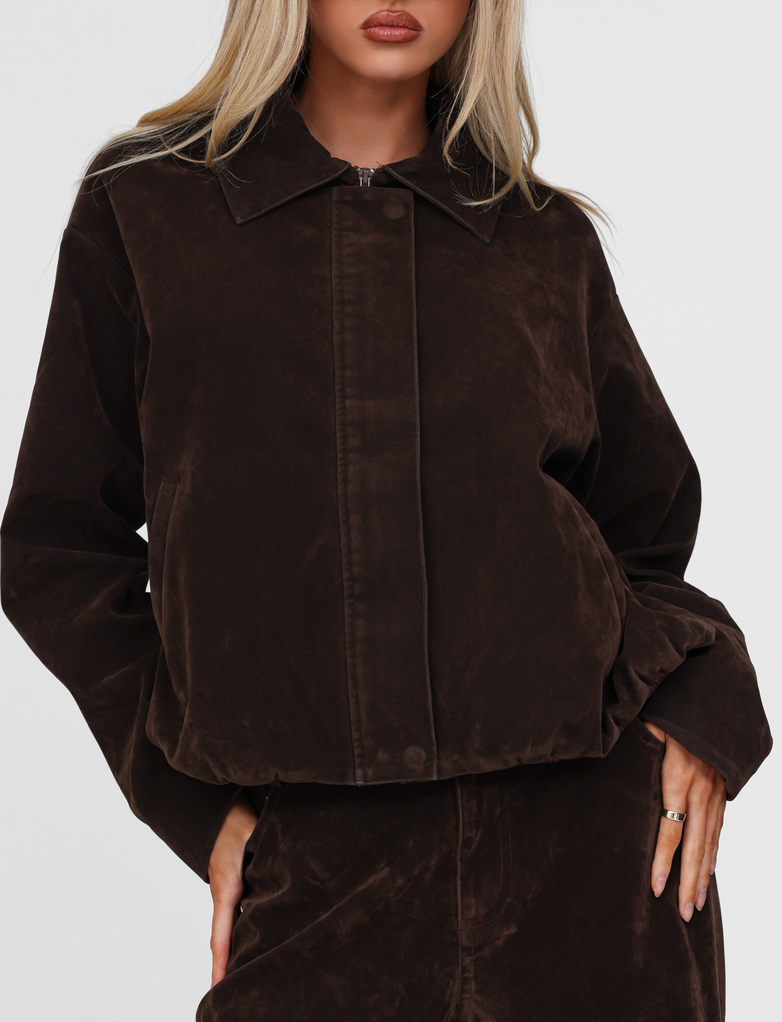A woman with long blonde hair wears the TIGERMIST CAMELLIA BOMBER JACKET in chocolate suede, paired with a matching skirt. She stands against a plain white background, hand on her hip.