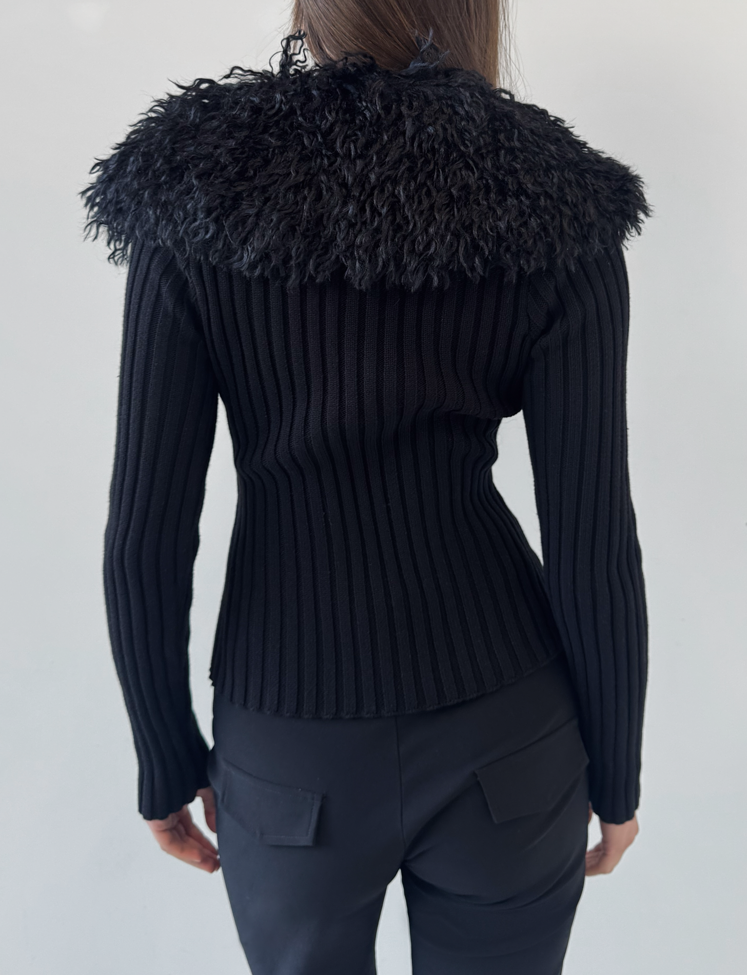 A person stands facing away, wearing the TIGERMIST BRISA JUMPER in black, which features a ribbed texture and a wide faux fur collar, paired with black pants with back pockets against a plain light background.