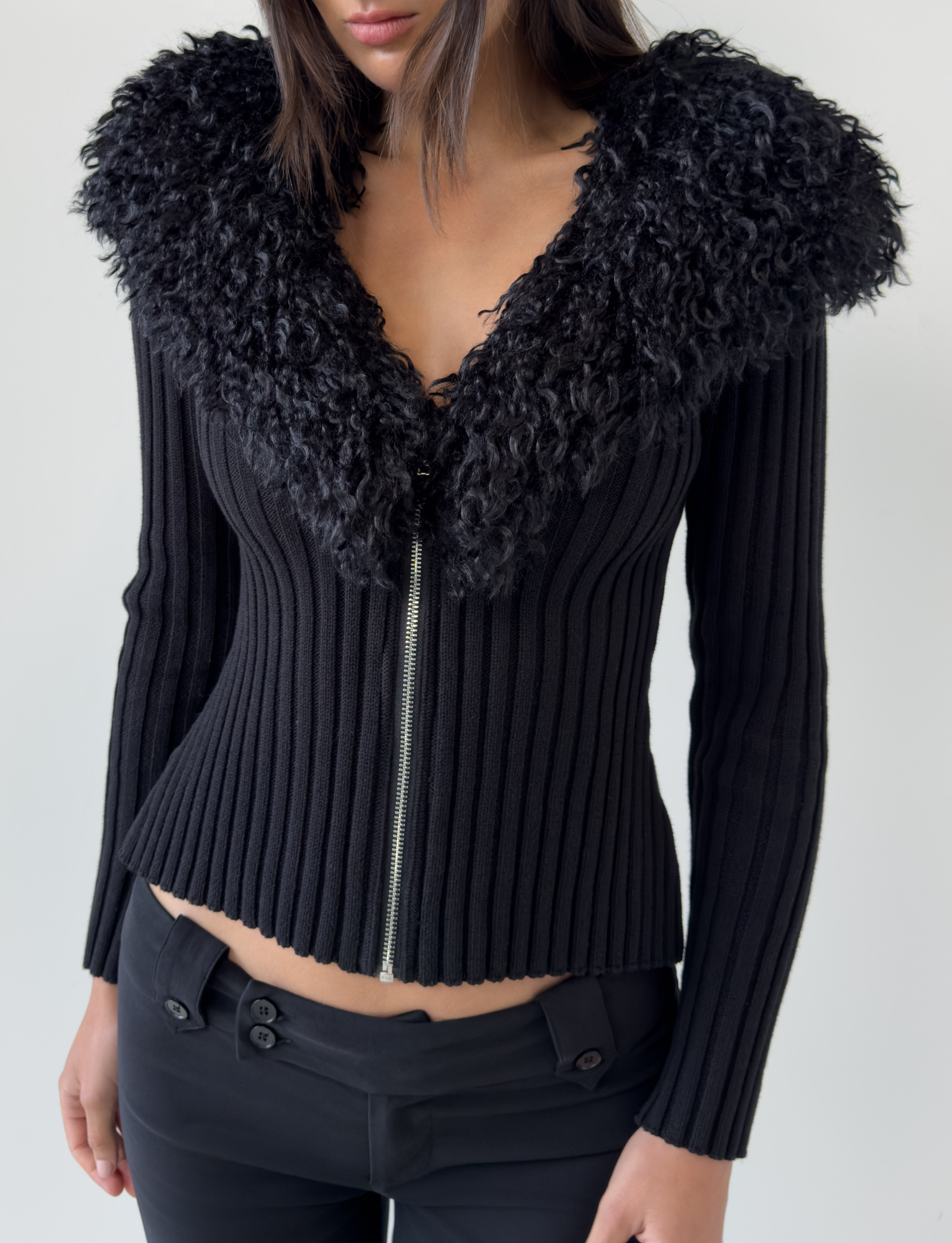 A woman wears the TIGERMIST BRISA JUMPER in black, a fitted ribbed jacket with a large faux fur collar and silver front zipper, paired with black pants. The jacket is zipped up; the photo is cropped at her head and mid-thigh.