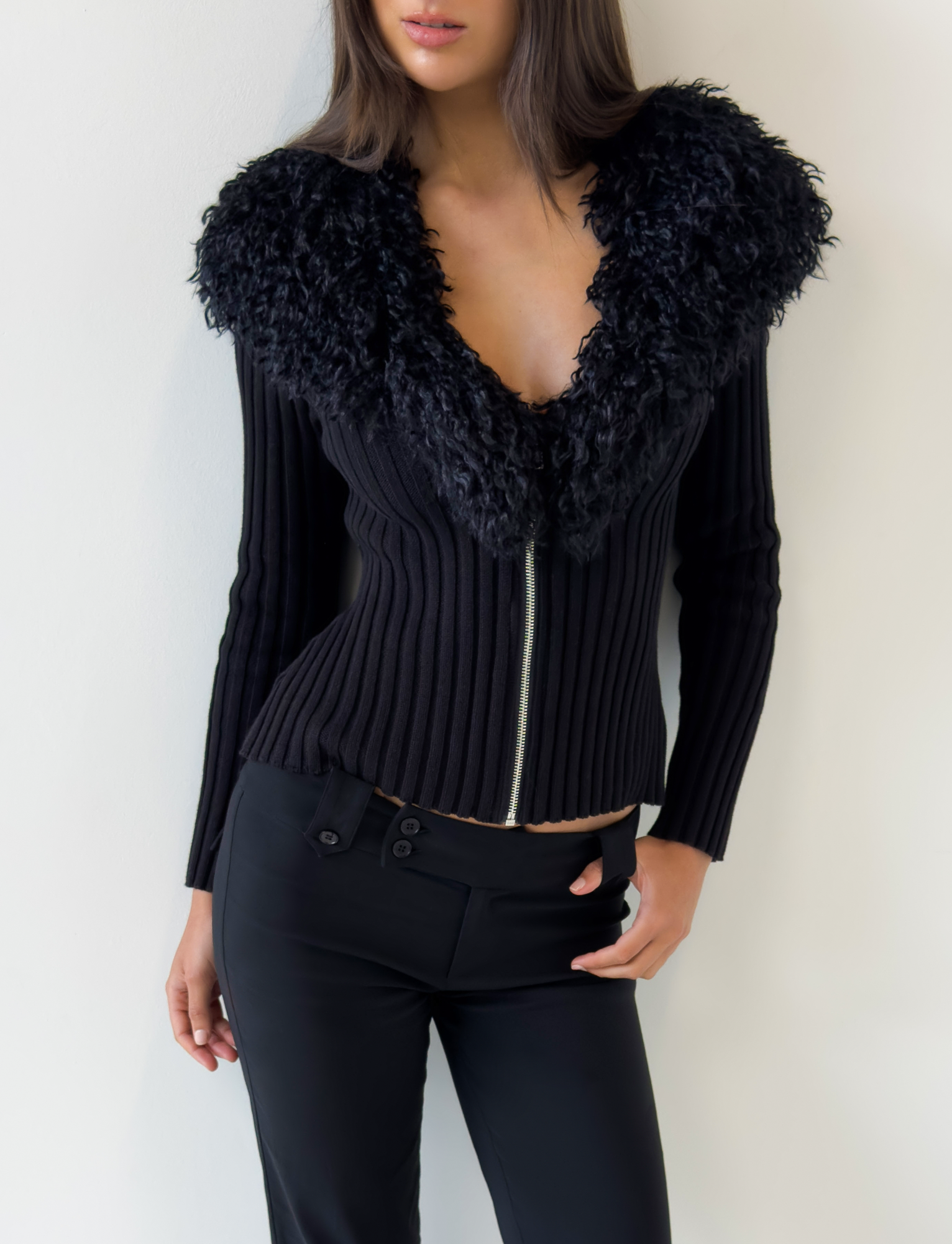 A woman models the TIGERMIST BRISA JUMPER - BLACK, featuring a ribbed design, large curly faux fur collar, and silver zipper. She pairs it with black pants and stands against a white wall, her face partially out of frame.