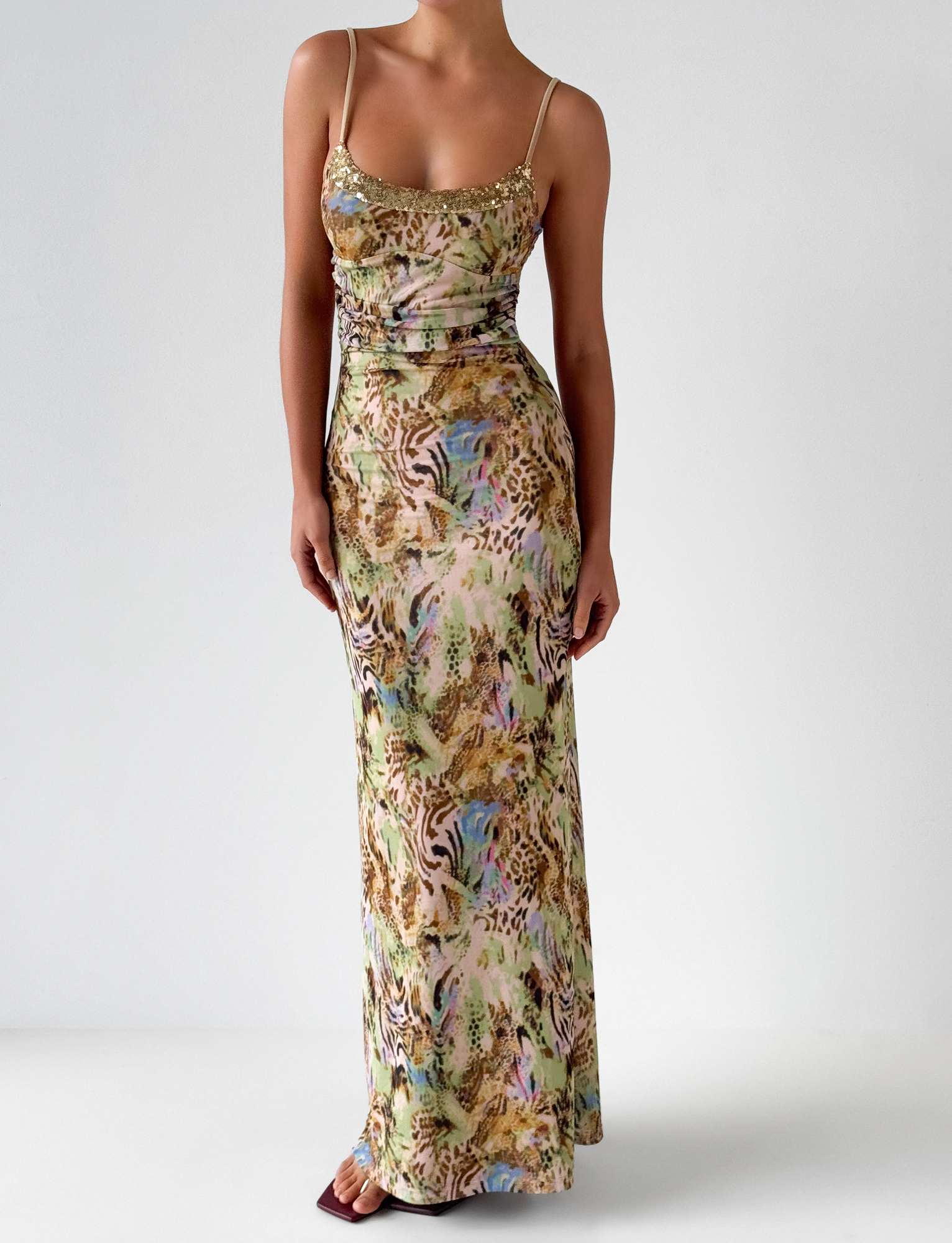 A woman poses against a plain background in the TIGERMIST ELYSIUM MAXI DRESS - ANIMAL SWIRL, a sleeveless, floor-length yellow mesh dress with thin straps and vibrant animal print. Her hands rest gently in front as she turns her head slightly.
