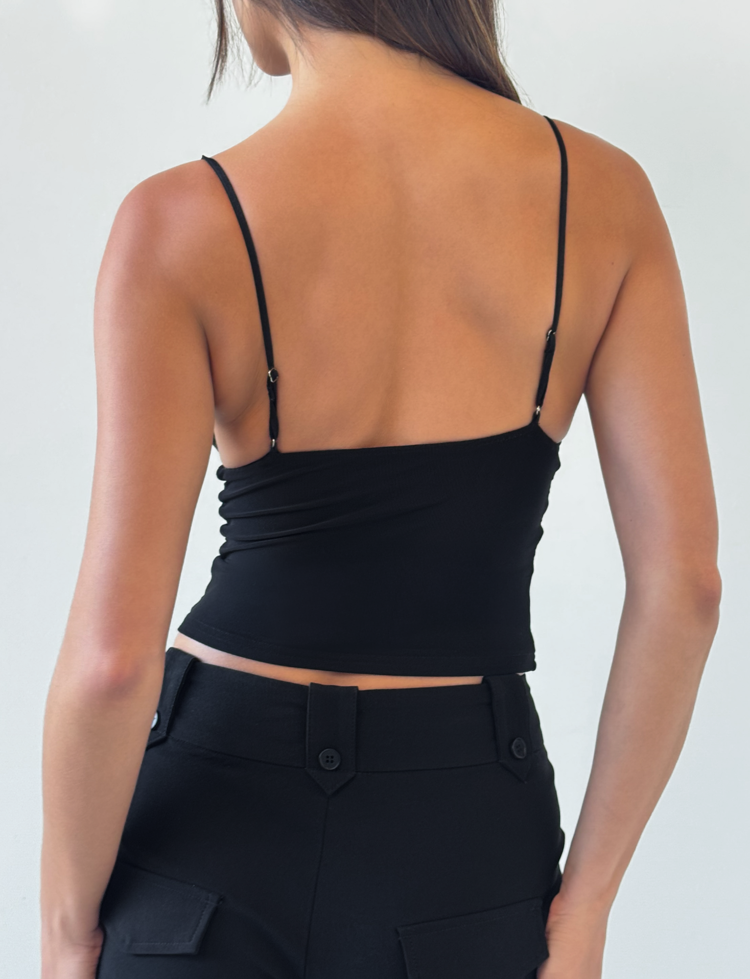A woman faces away, wearing the TIGERMIST FENELLA TOP - BLACK, a sequin mesh tank with adjustable spaghetti straps, paired with high-waisted black pants. Her loose brown hair contrasts against a plain light background.