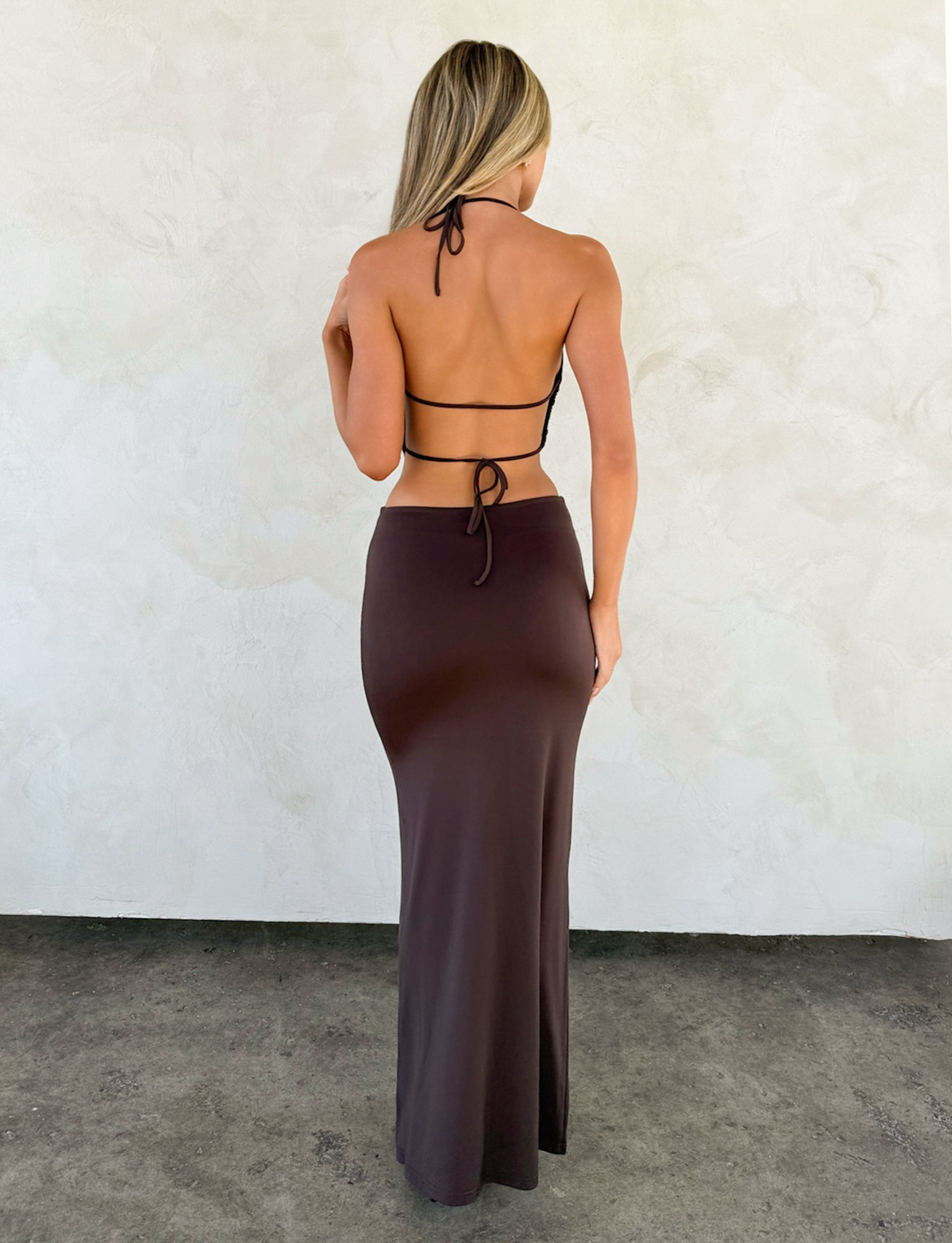 A woman with long blonde hair faces a light textured wall, wearing the TIGERMIST SURIYA MAXI DRESS in chocolate brown—a backless cowl neck style with adjustable halter straps and a gold waist chain, standing on a dark concrete floor.