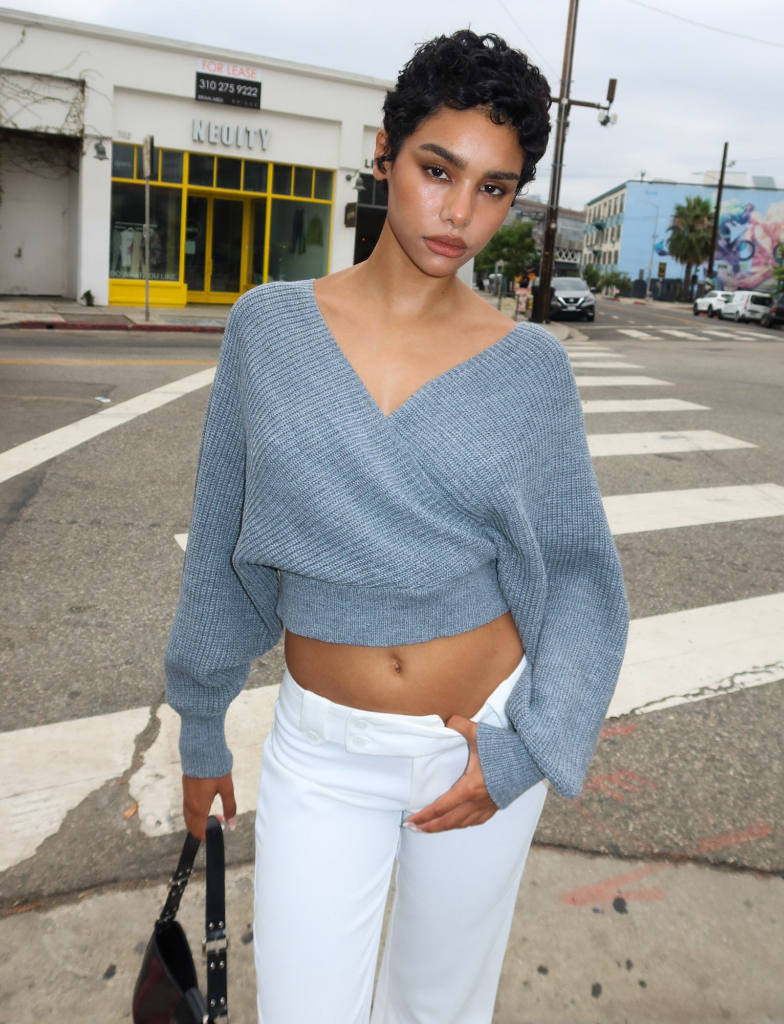 A person with short curly hair stands on a city sidewalk wearing the TIGERMIST RAVEN JUMPER in grey and white pants, holding a black handbag and looking at the camera with buildings and a crosswalk behind them.