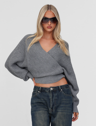 A person with long blonde hair wears large dark sunglasses, blue jeans, and the TIGERMIST RAVEN JUMPER - GREY, featuring bell sleeves, standing against a plain light background with one hand on their hip.