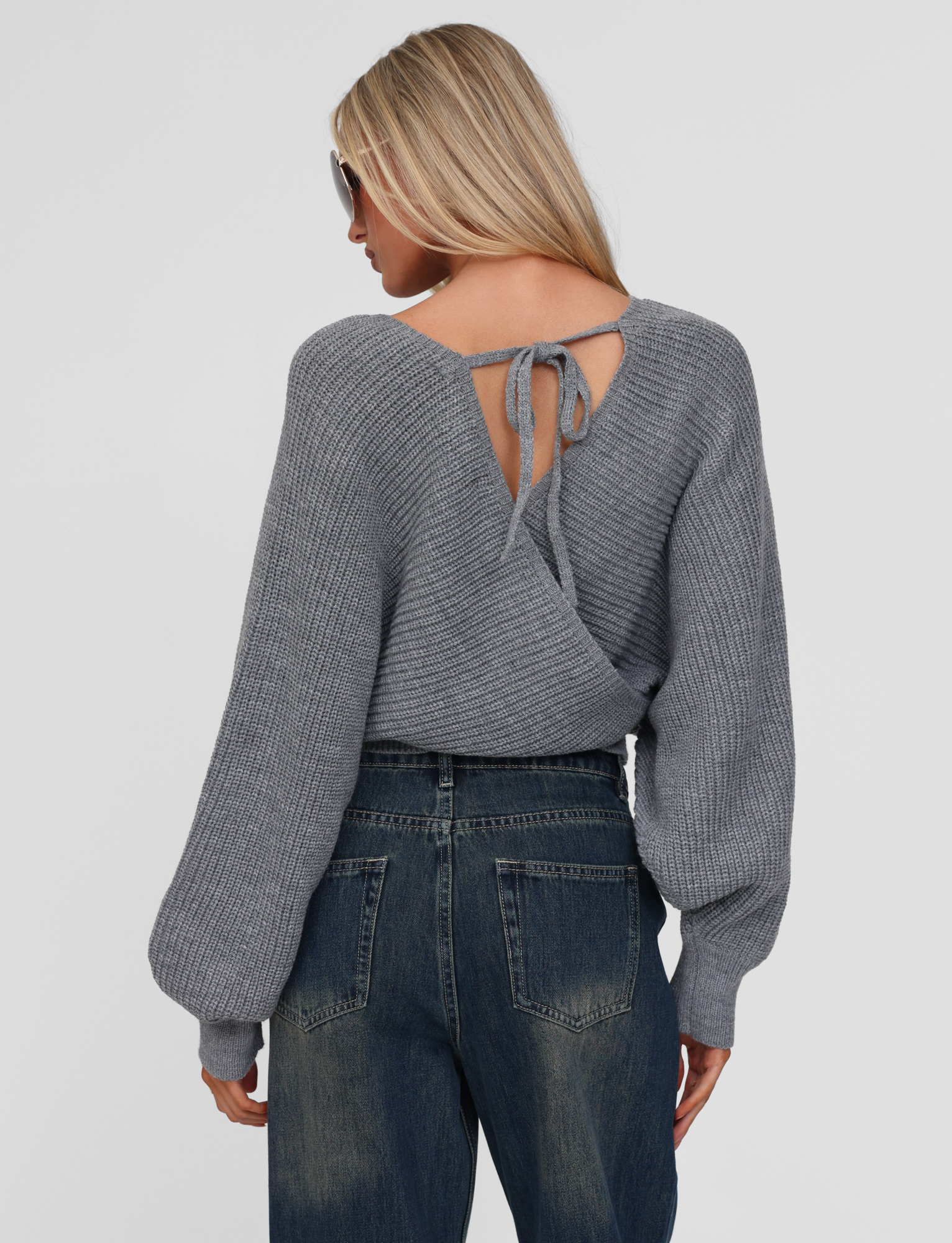 A woman with long blonde hair and sunglasses wears the TIGERMIST RAVEN JUMPER - GREY, featuring a crisscross back, tie detail, and bell sleeves, paired with high-waisted blue jeans. She faces away from the camera.