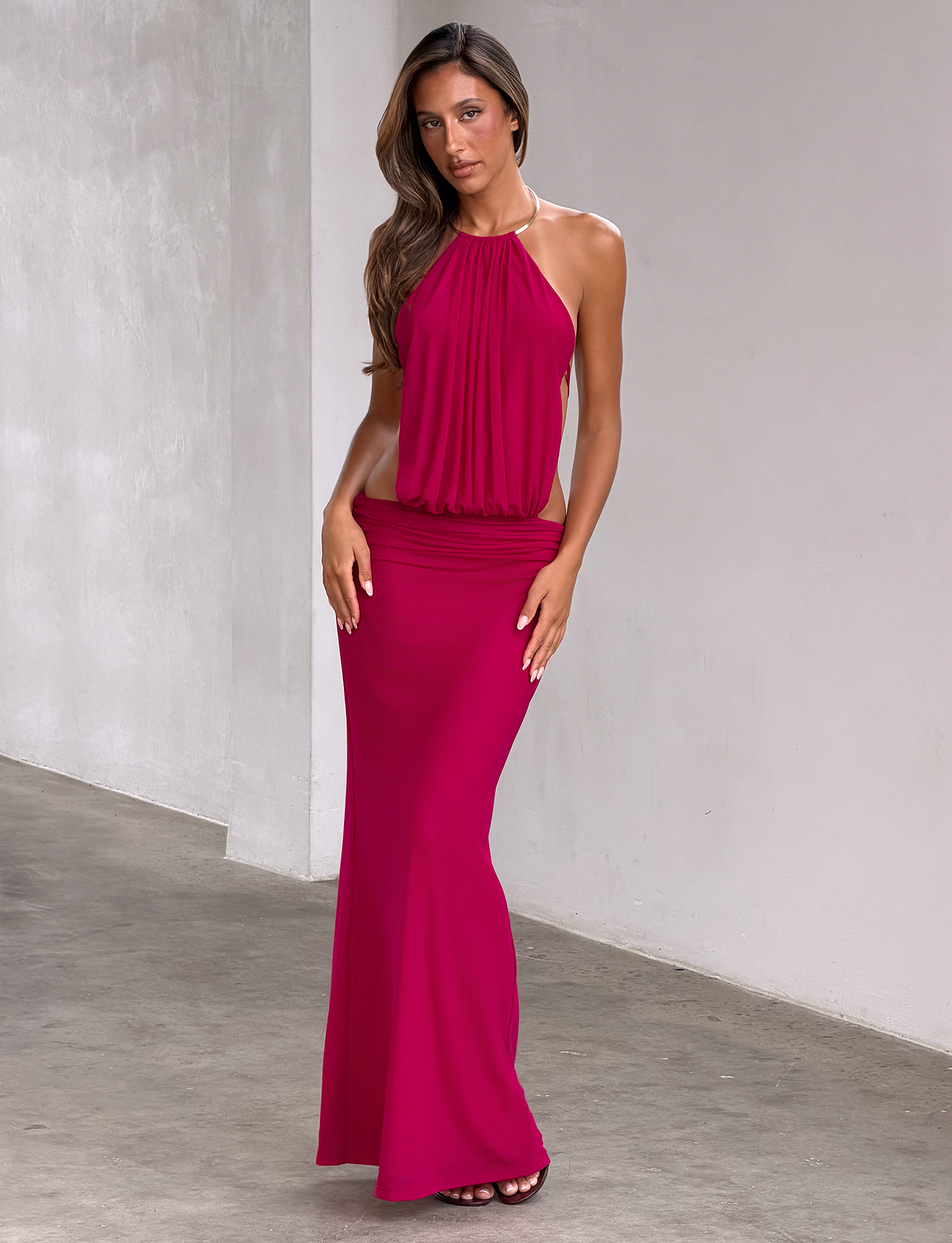 A woman with long, wavy brown hair poses indoors against a plain light wall and concrete floor, wearing the TIGERMIST EVANNA MAXI DRESS in berry—a sleeveless, floor-length mesh gown featuring a halter neck and gold-trimmed neckline.