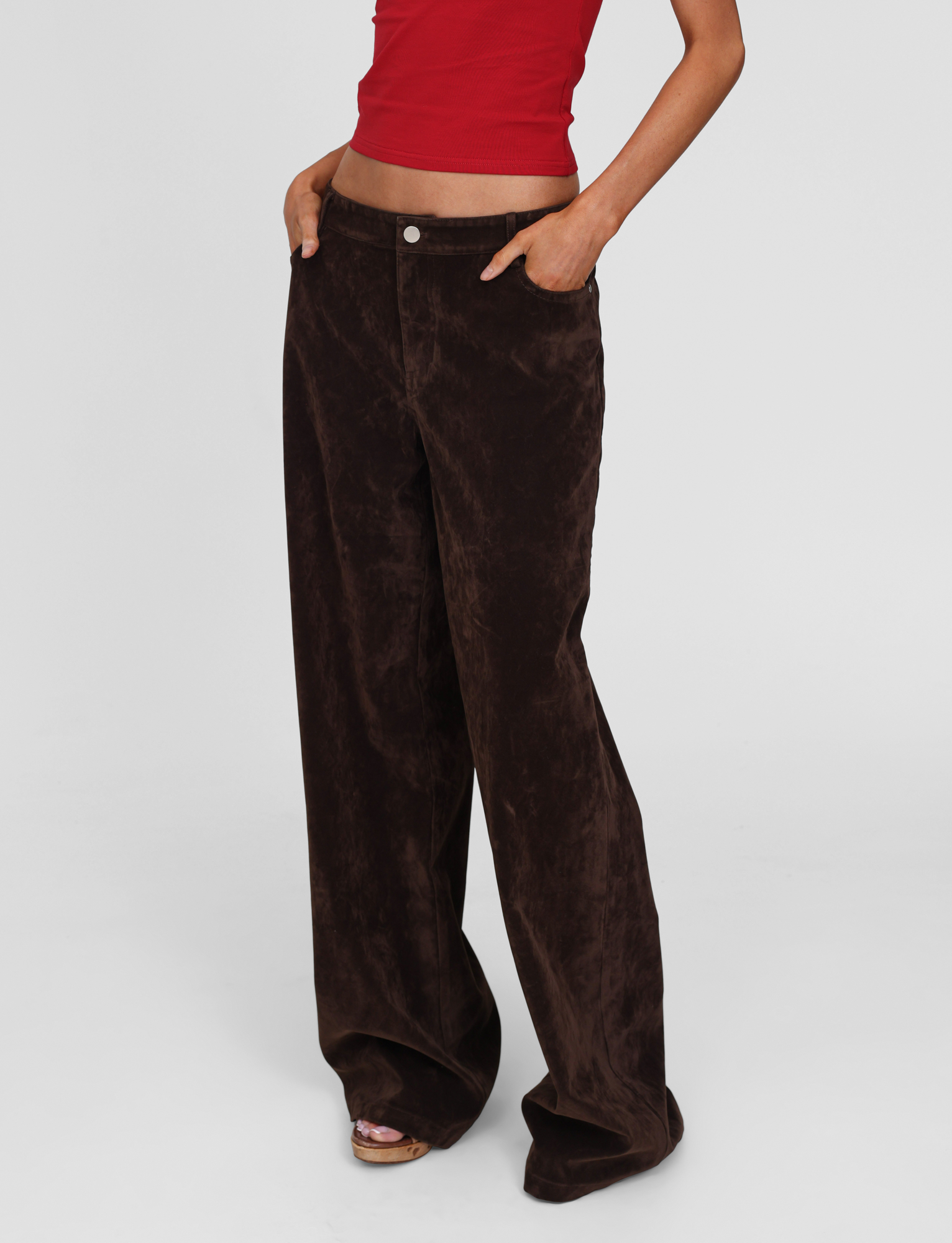 A person models the TIGERMIST CAMELLIA PANT in chocolate suede, paired with a red crop top. Their hands are in their pockets and their head is not visible, standing against a plain white background.