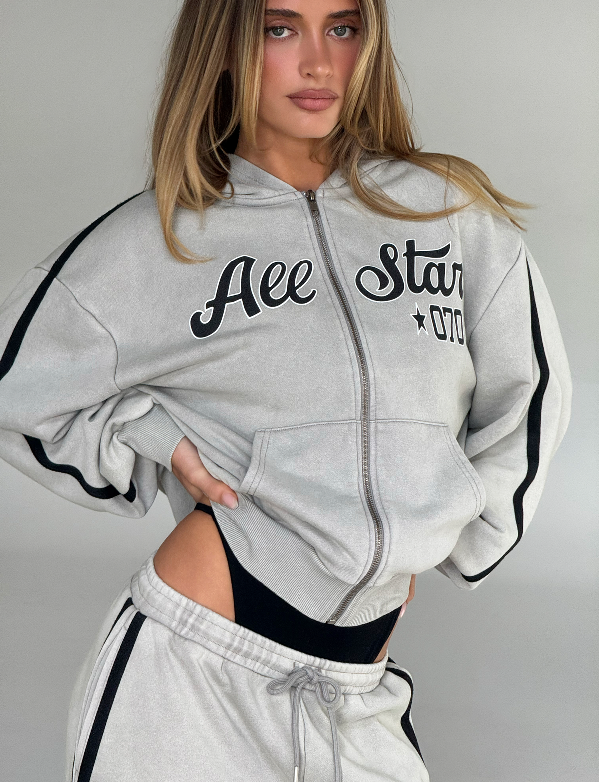 A woman poses with one hand on her hip, wearing the TIGERMIST PAYTON HOODIE - STONE and matching sweatpants, slightly revealing black undergarments above the waistband.
