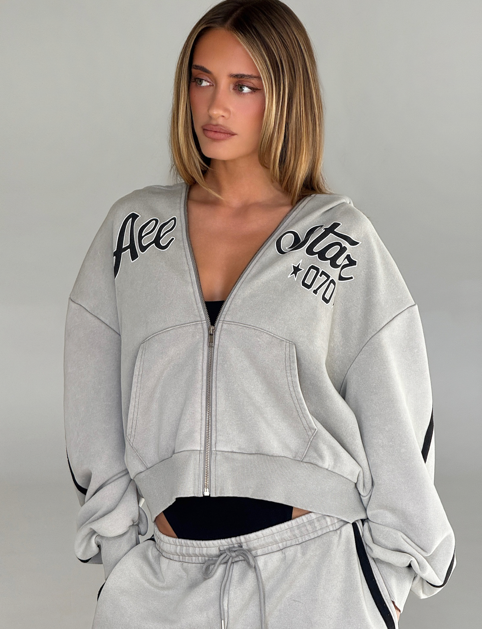 A young woman with straight, light brown hair wears the TIGERMIST PAYTON HOODIE in Stone with black lettering and matching sweatpants, gazing to the side with a neutral expression against a plain background.