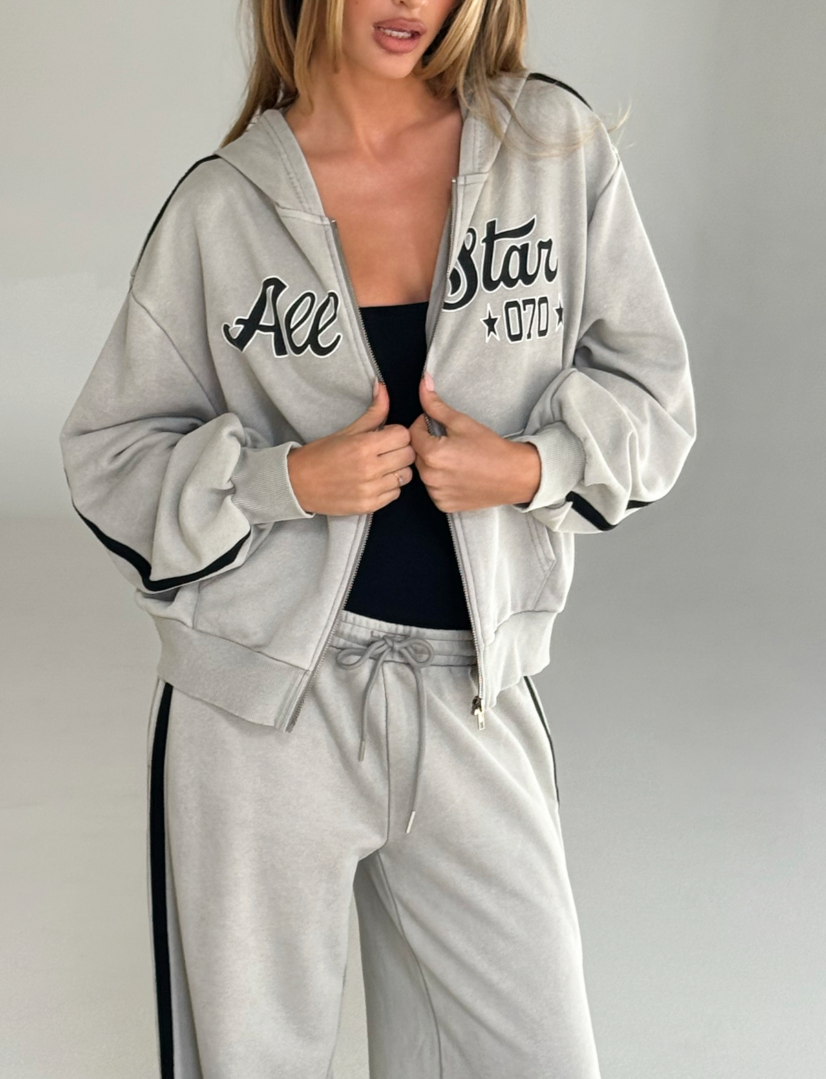 A woman wears the TIGERMIST PAYTON HOODIE in Stone, a vintage-inspired lightweight terry hoodie, partially open over a black top, paired with matching sweatpants.