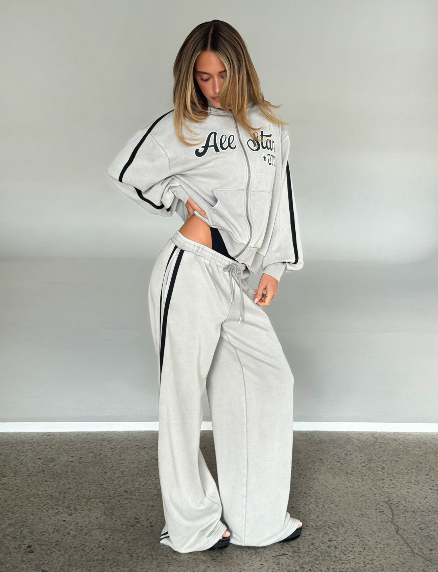 A woman models the TIGERMIST PAYTON TRACKPANT - STONE, loose-fitting with black stripes, standing indoors on a concrete floor against a plain light gray background that highlights the sporty, relaxed vibe of these trackpants.
