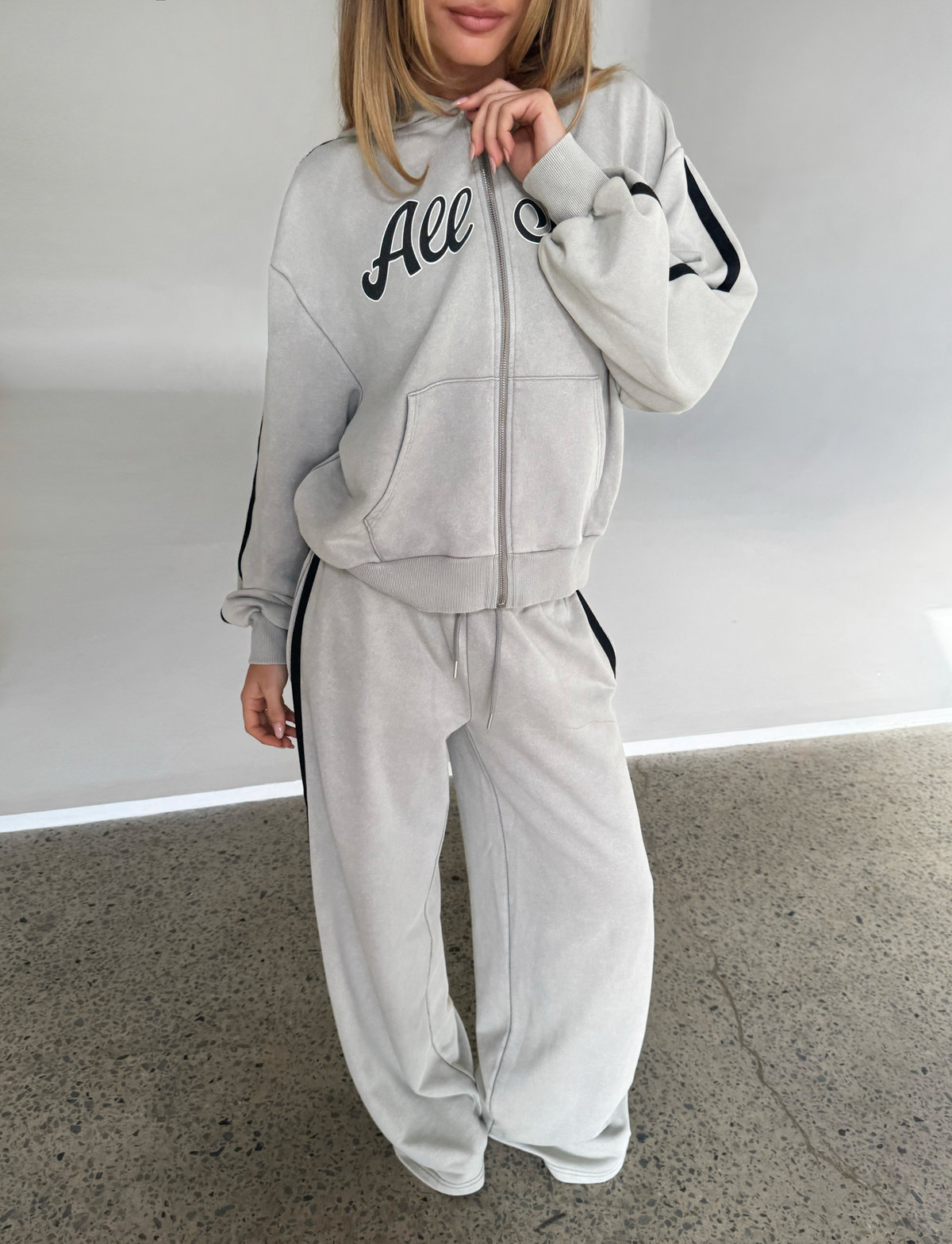 A woman stands indoors wearing TIGERMIST's PAYTON TRACKPANT - STONE, a light gray sporty cotton trackpant with black stripes, paired with a matching zip-up hoodie featuring "All" on the chest. Her face is partially out of frame.
