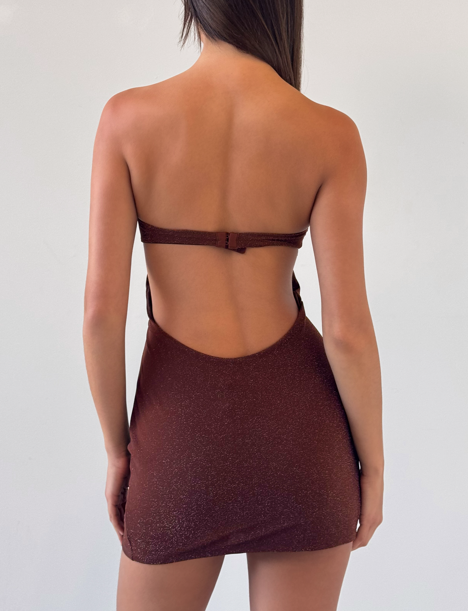 A woman faces away from the camera, modeling the TIGERMIST KESTREL MINI DRESS in Chocolate—a shimmering, fitted dress with thin straps—against a plain light background.