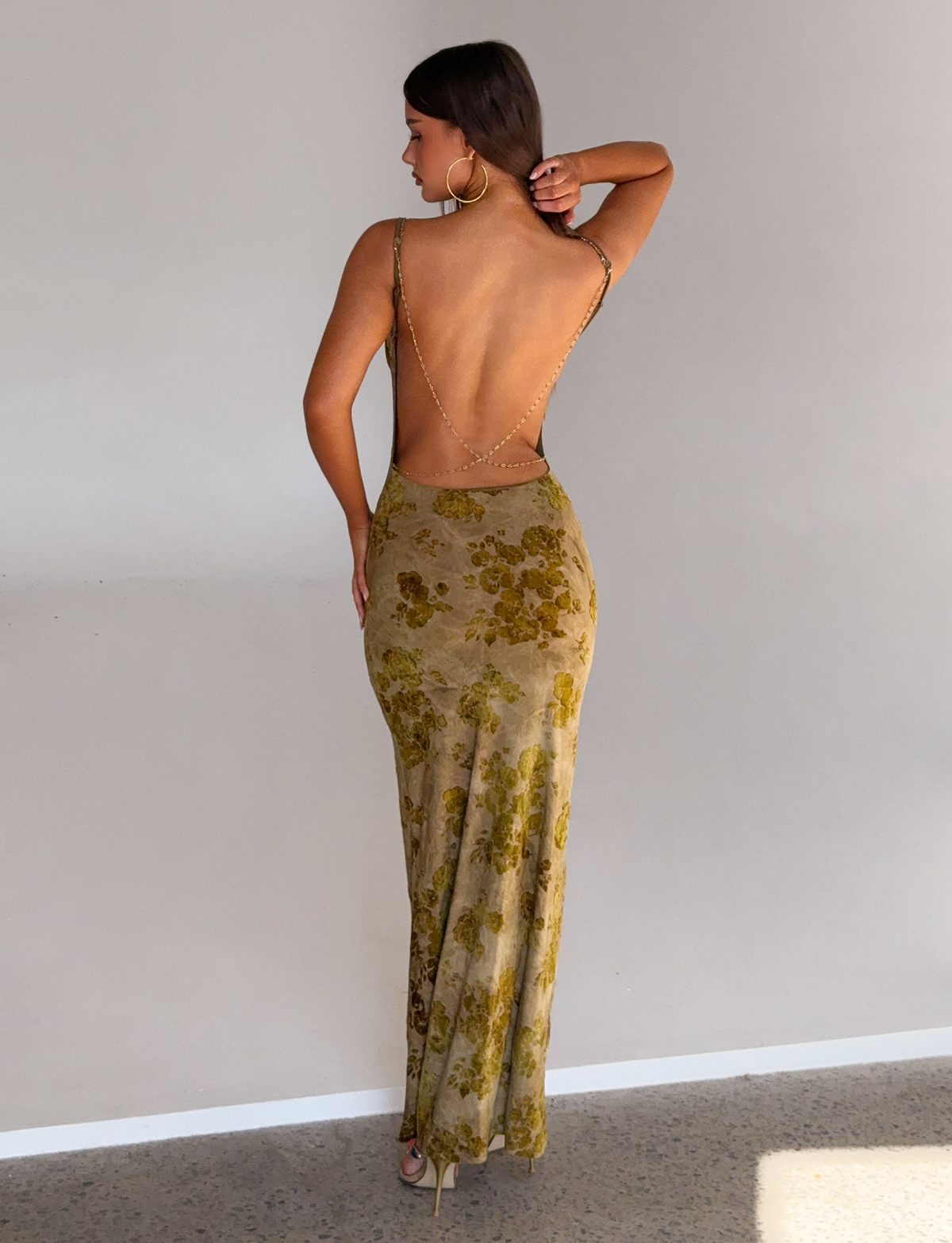 A woman stands with her back to the camera, wearing the TIGERMIST SPARROW MAXI DRESS in Green Burnout—a form-fitting, floor-length dress with gold chain straps and a yellow floral pattern—touching her hair against a plain light wall.