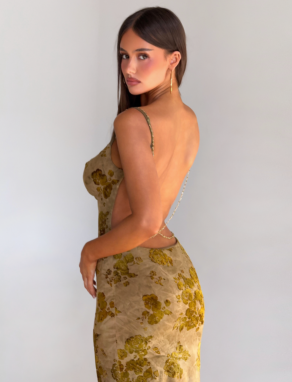 A woman with long brown hair models the TIGERMIST SPARROW MAXI DRESS - GREEN BURNOUT, a backless floral dress with delicate beaded chain straps, standing sideways and glancing over her shoulder against a plain light background.