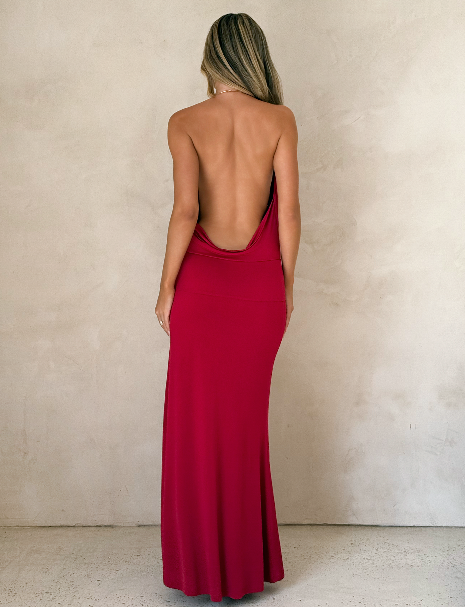 A woman with long blonde hair faces a textured beige wall, wearing the TIGERMIST TARRIN MAXI DRESS in red, featuring a floor-length silhouette, cowl neckline, daring thigh-high slit, and low draped back.