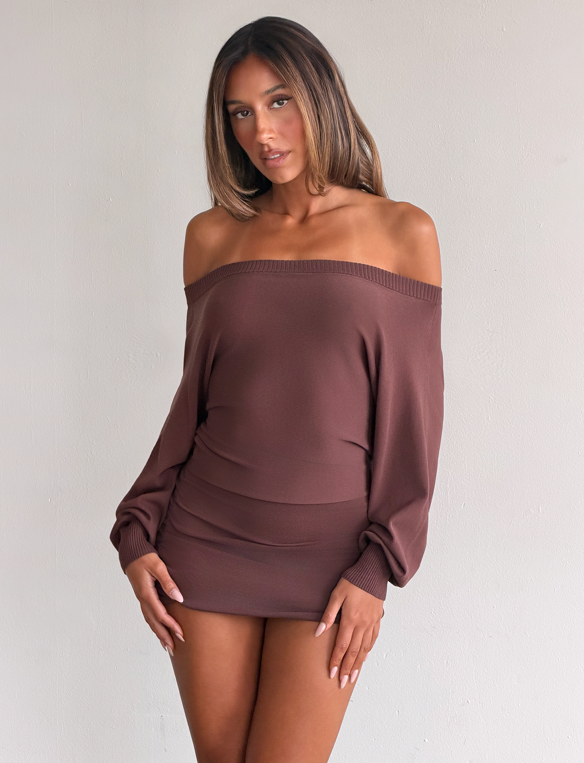 A woman poses against a plain light wall in the TIGERMIST JANINE MINI DRESS - CHOCOLATE, an off-shoulder mini with a ruched waist. Her straight, shoulder-length hair and confident expression complete the look.