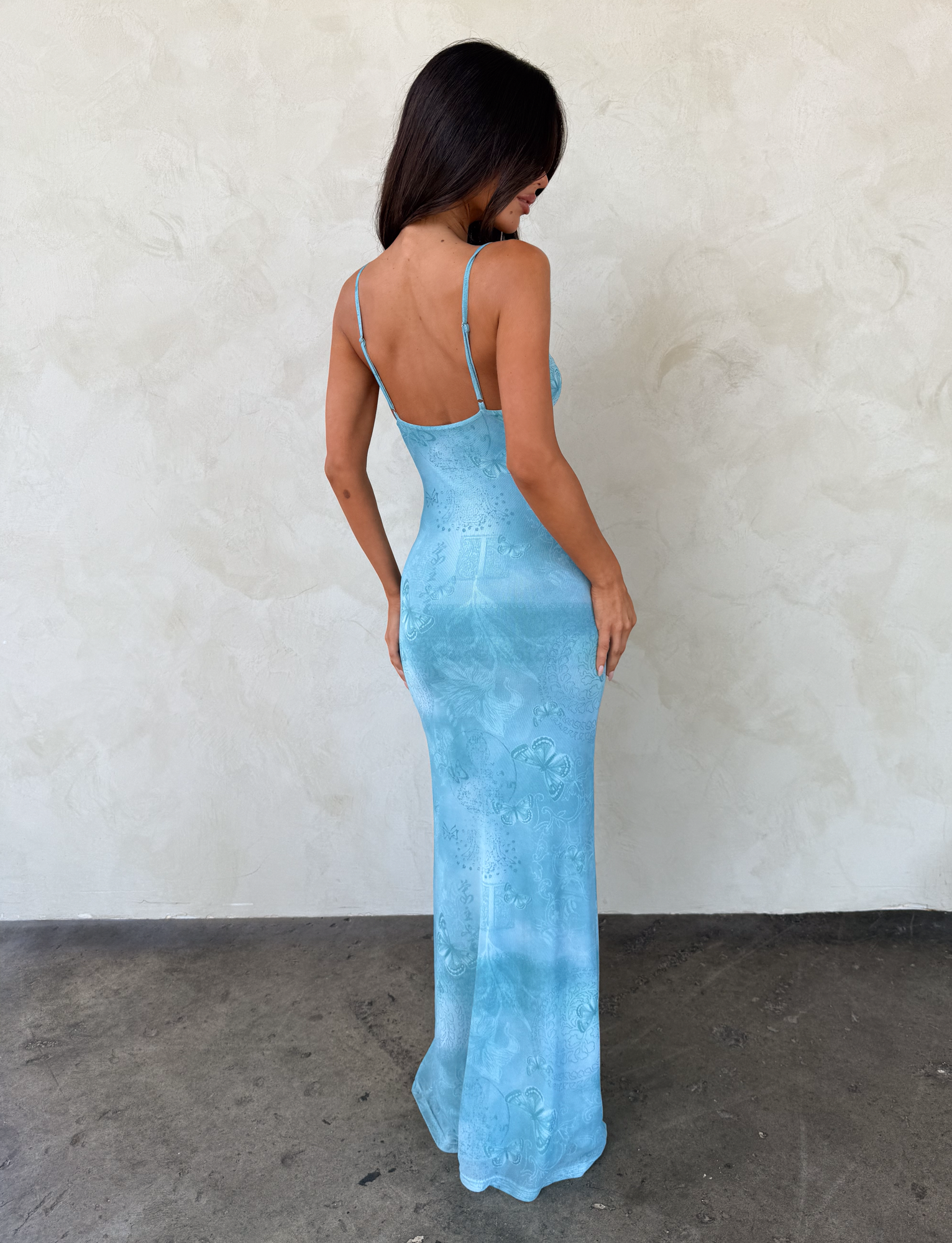A woman with long dark hair faces away, wearing the TIGERMIST GARNET MAXI DRESS in Teal Butterfly Paisley, featuring adjustable straps, against an off-white textured wall and concrete floor.