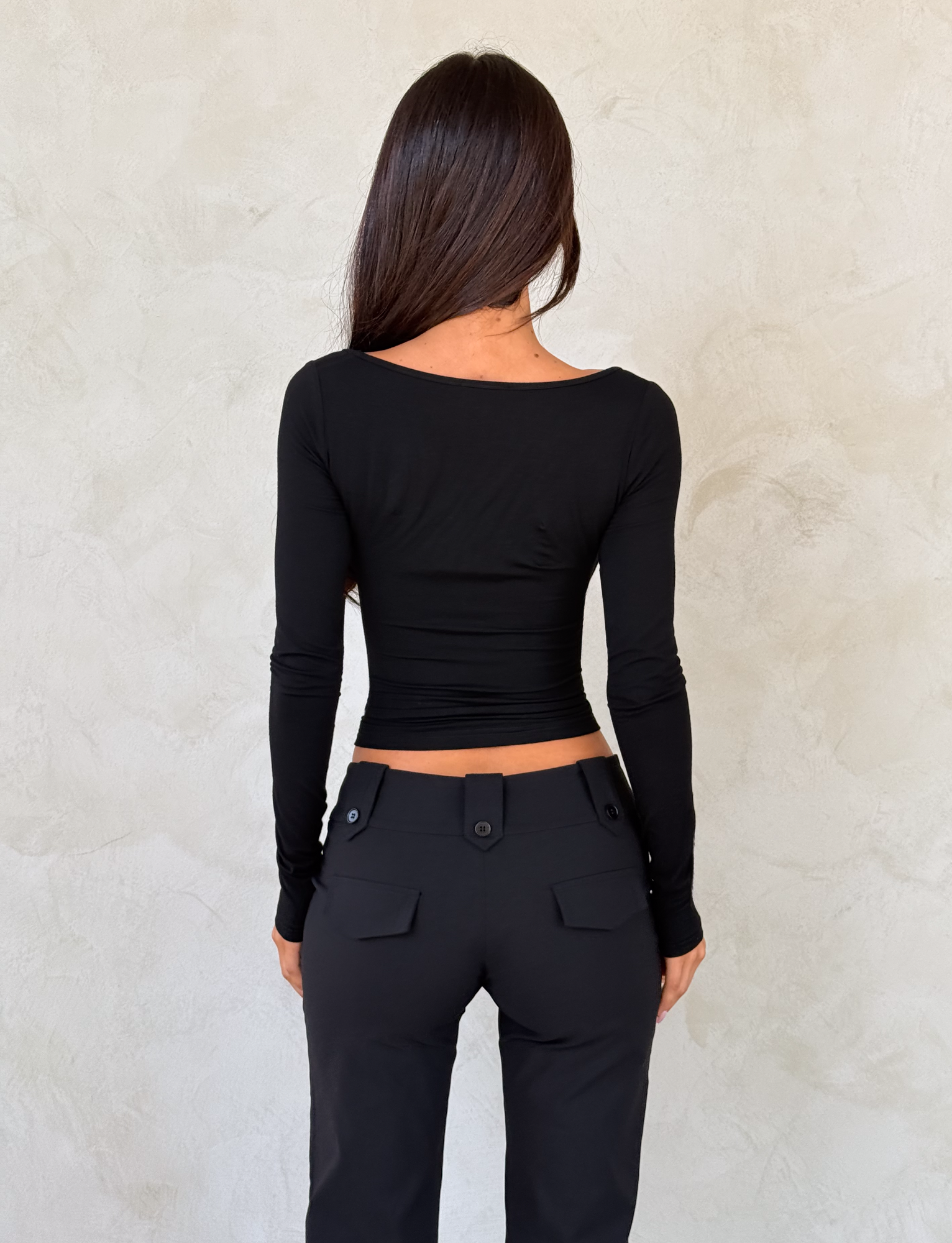 A woman with long dark hair, seen from behind, wears the TIGERMIST DAMELA LONG SLEEVE TOP in black with an adjustable neckline and fitted black pants featuring buttoned pockets, standing against a light textured wall.