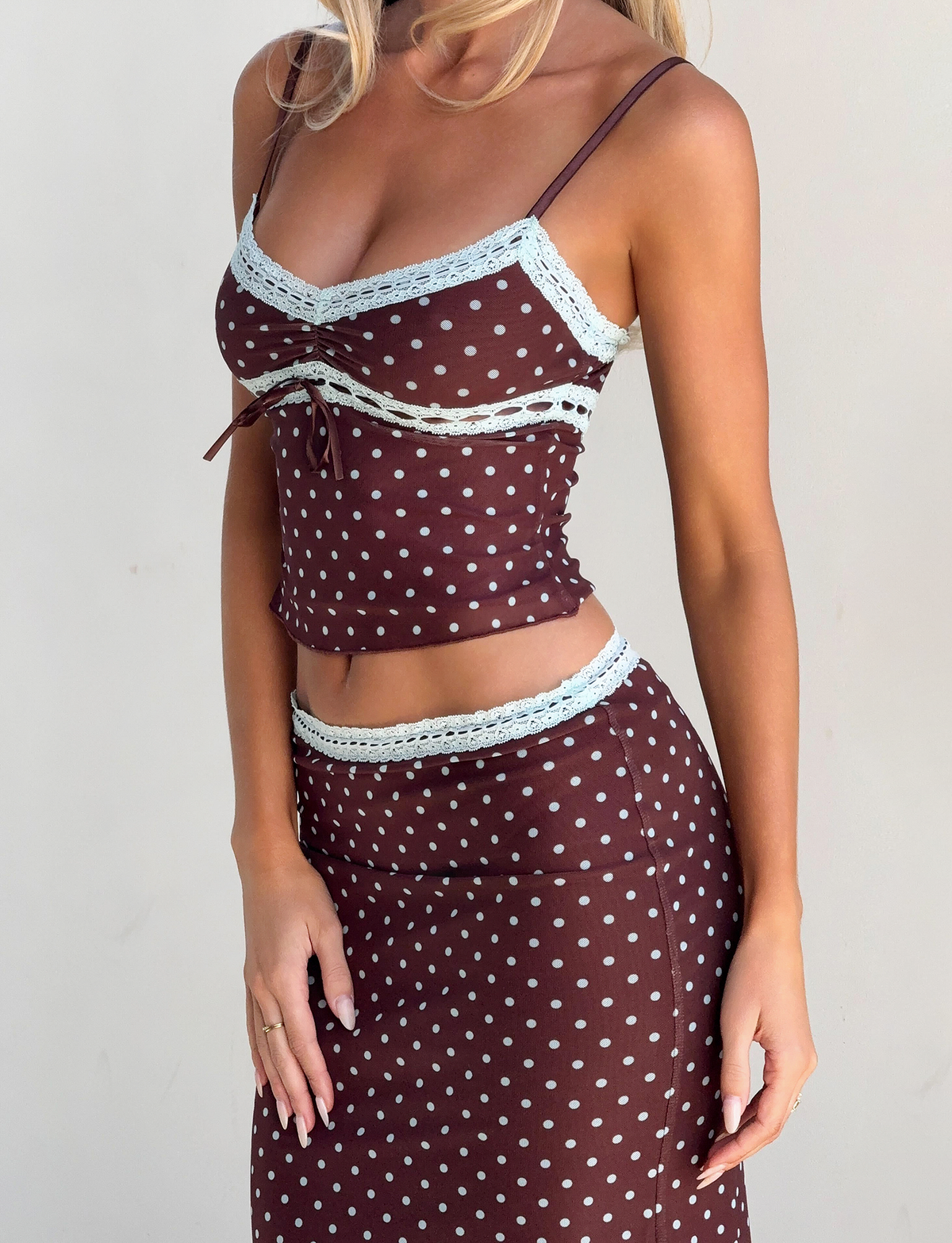 A woman models the TIGERMIST AMARA TOP - POLKA DOT, a brown sleeveless crop top with white lace trim, paired with a matching skirt. She stands against a plain light background, and her face is not visible.