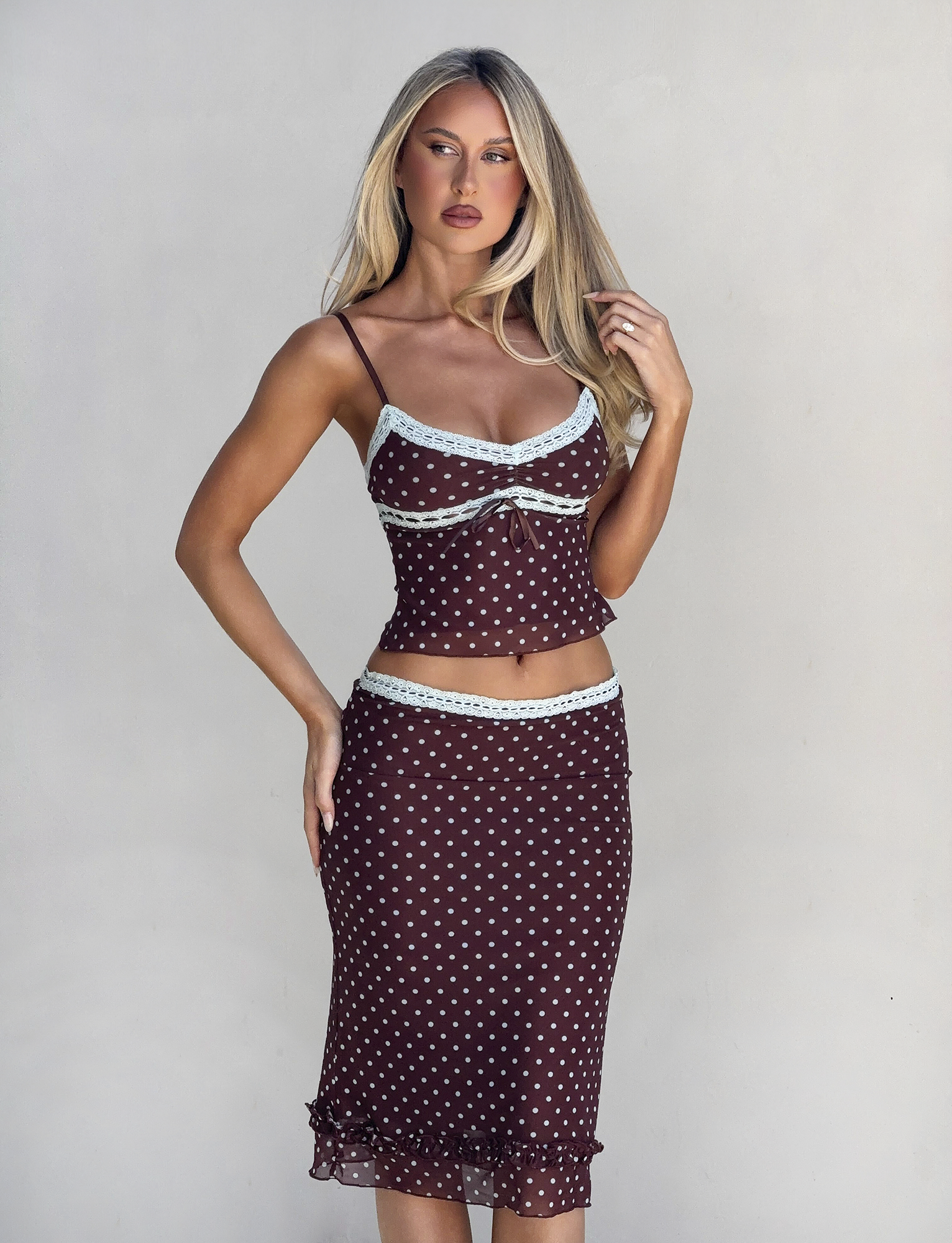 A woman with long blonde hair poses against a light background, wearing the TIGERMIST AMARA MIDI SKIRT - POLKA DOT and matching spaghetti strap top, both brown with white dots and light blue lace trim.