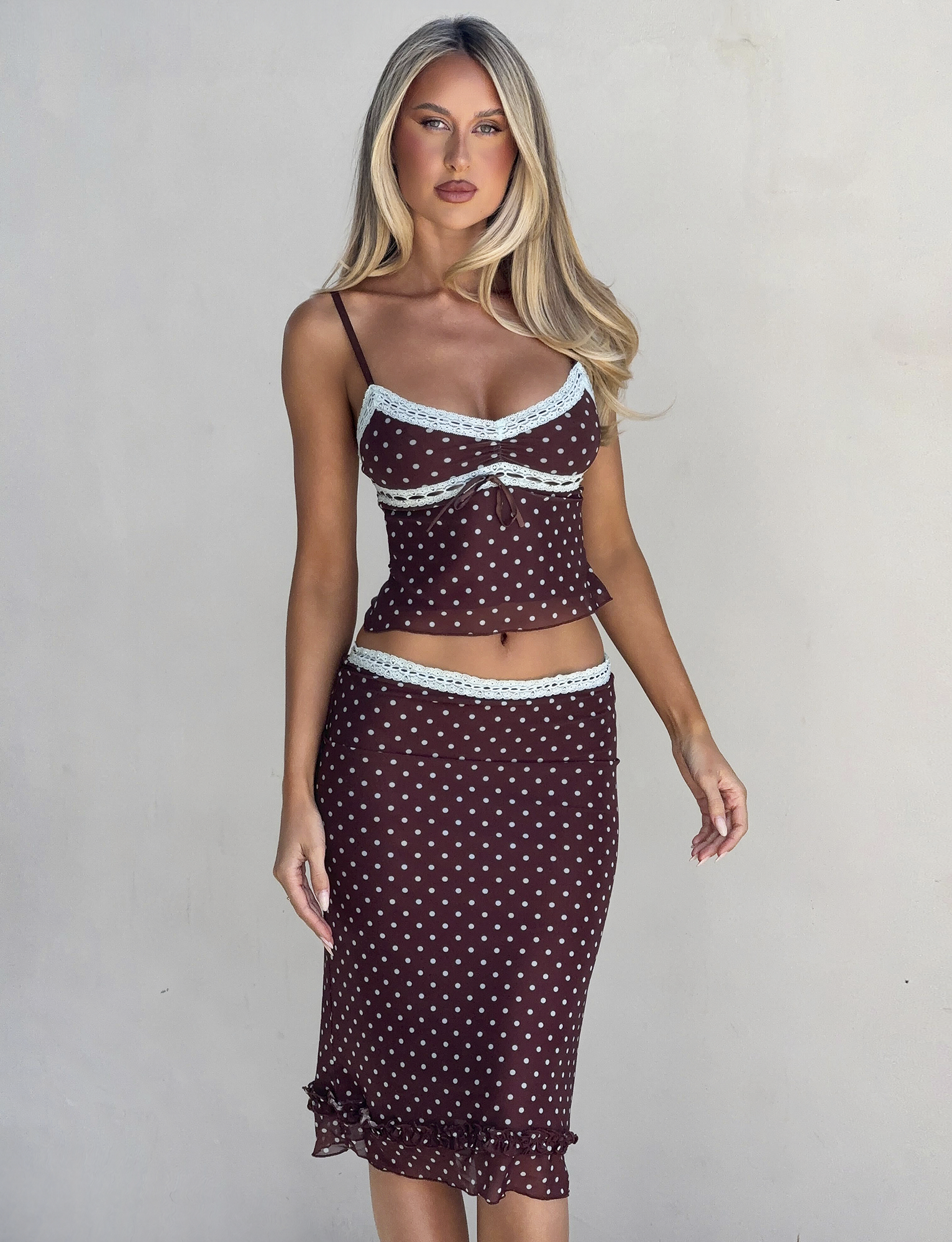 A woman with long blonde hair models the TIGERMIST AMARA MIDI SKIRT - POLKA DOT, a brown two-piece outfit with light blue trim and spaghetti straps, against a plain light background.