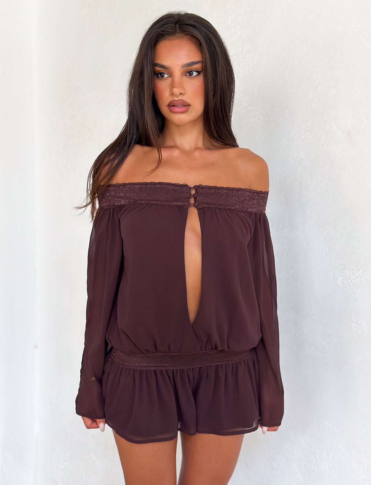 Woman with long dark hair wears the TIGERMIST TESSIE PLAYSUIT in brown, featuring off-shoulder style, ruffled mini shorts, and a front keyhole cutout, standing against a plain white background.