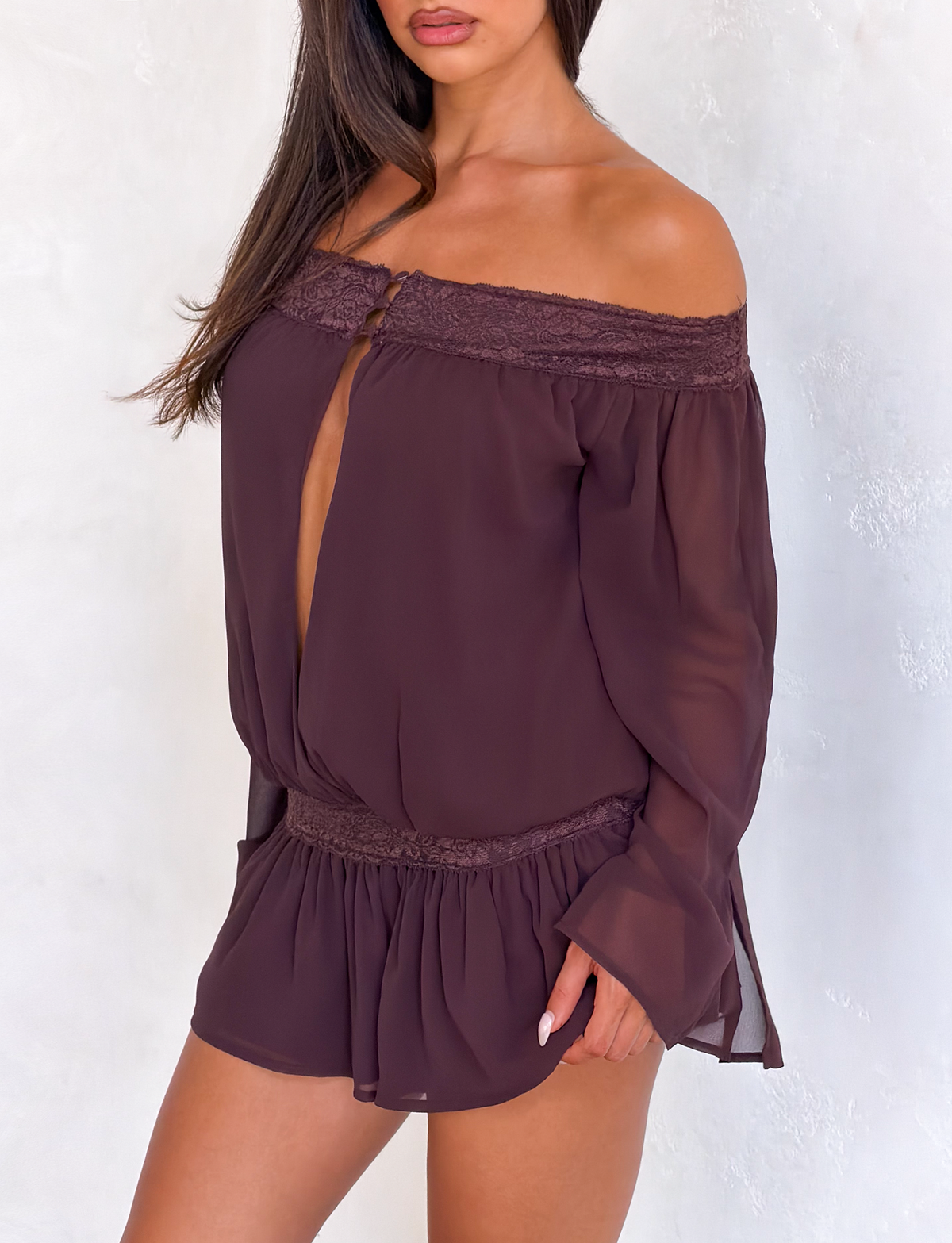 A woman models the TIGERMIST TESSIE PLAYSUIT in brown—an off-shoulder, long-sleeve romper with lace trim and ruffled mini shorts—posing against a light textured background with her hair down, face partially out of frame.