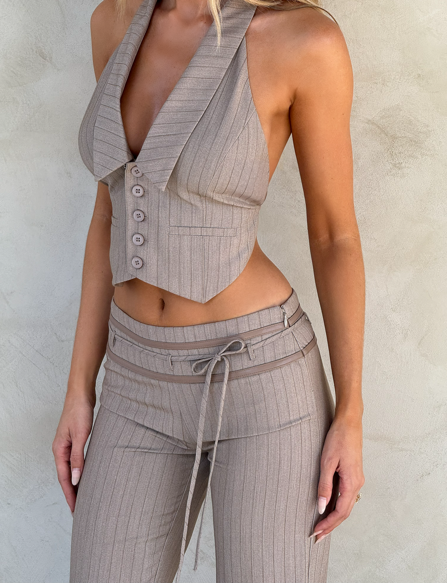 A woman models the TIGERMIST FEDRA VEST - TAN PINSTRIPE, a sleeveless vest with wide lapels, paired with matching high-waisted, tie-belt pants, standing against a light textured wall with her face unseen.