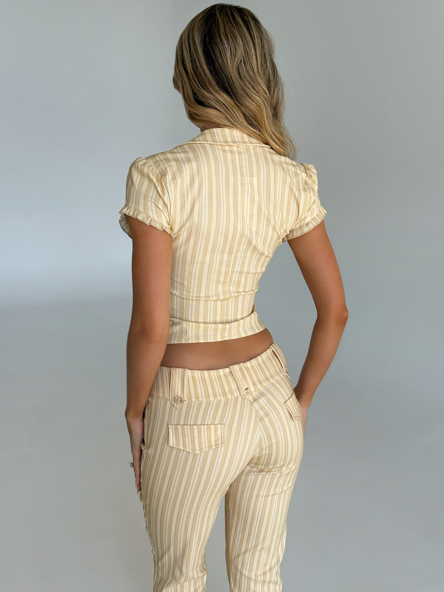 A woman with wavy blonde hair faces away in the fitted, cropped-length KITTIE CAPRI - YELLOW STRIPE set by TIGERMIST, featuring beige and white vertical stripes in soft stretch suiting, against a plain light background.