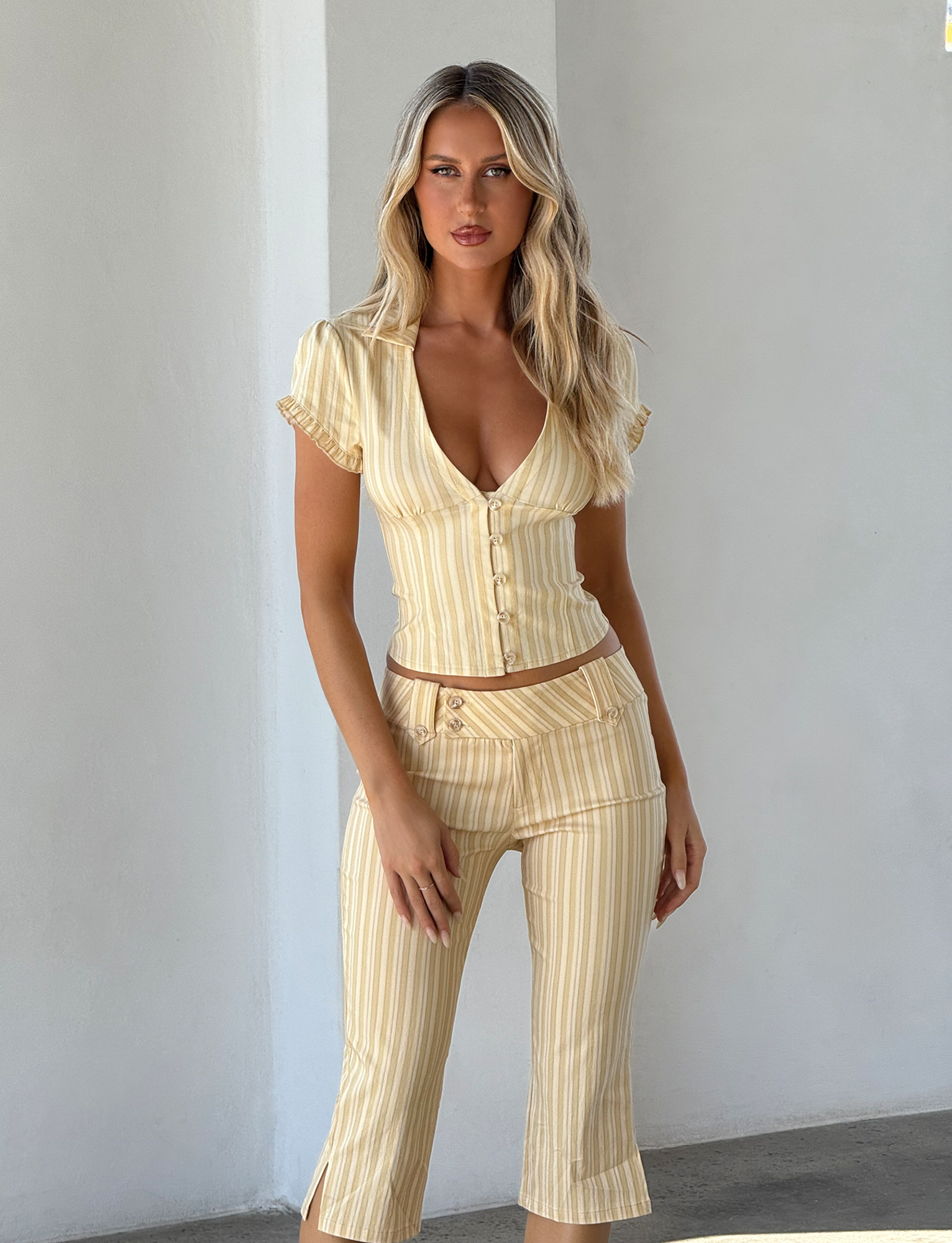 A woman with long blonde hair wears the TIGERMIST KITTIE SHIRT in yellow stripe, a fitted short-sleeved button-up, paired with matching cropped pants. She stands confidently against a light background.
