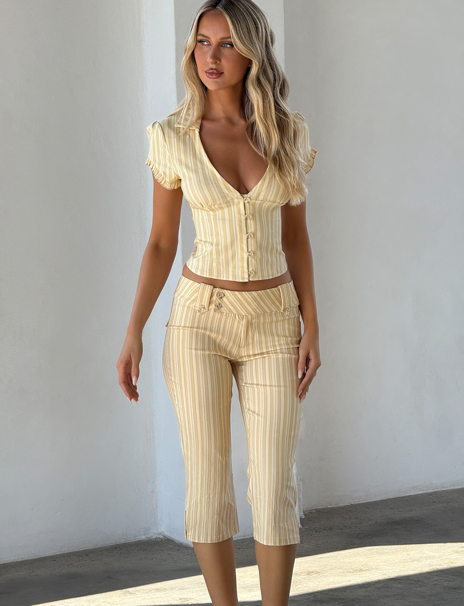A woman with long blonde hair models the TIGERMIST KITTIE CAPRI - YELLOW STRIPE set, featuring a fitted short-sleeve button-up top and matching high-waisted, cropped capri pants in yellow and white stripes against a white wall.