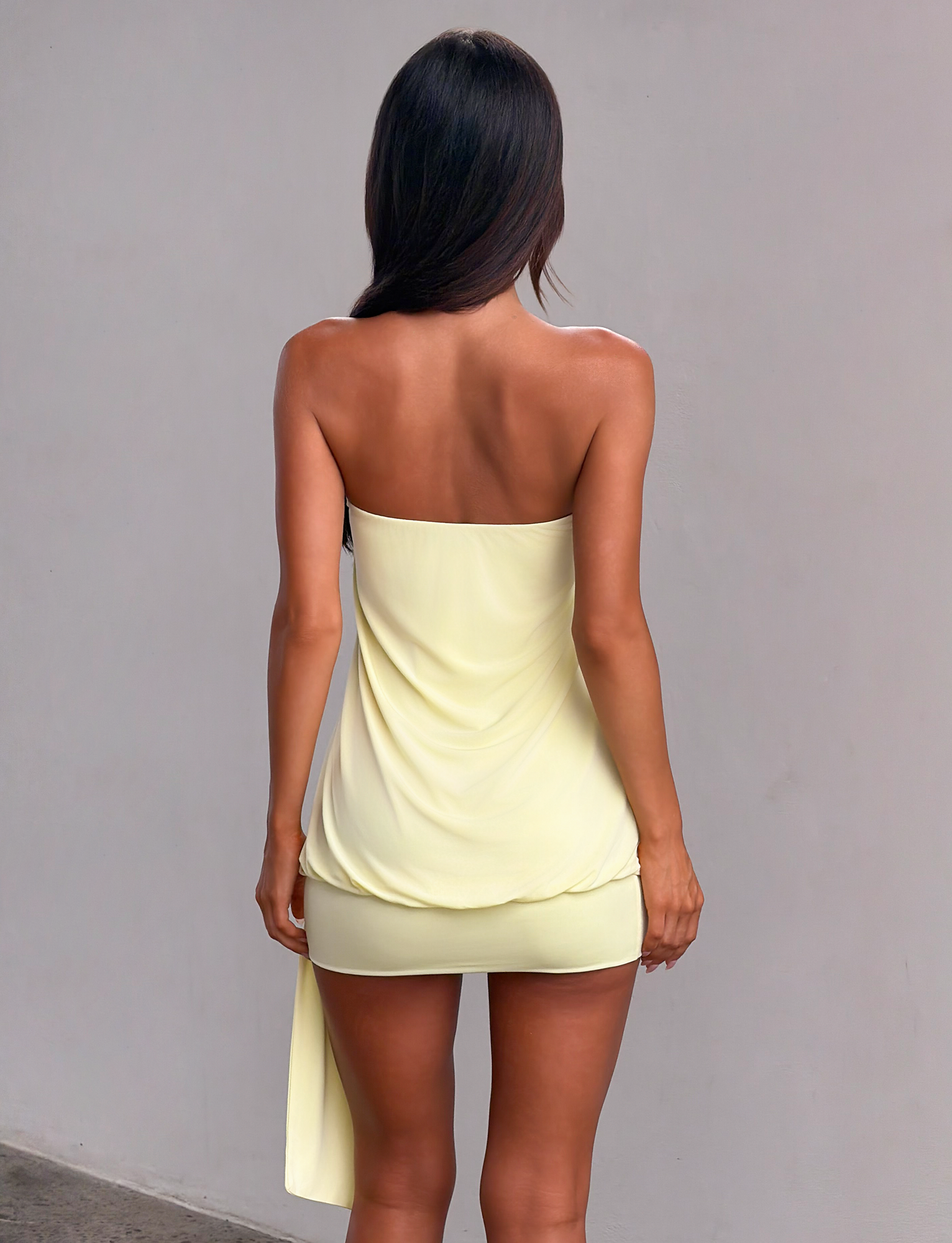 A woman with long dark hair stands facing away, wearing the TIGERMIST RAVENNA MINI DRESS in butter yellow, featuring a strapless design, draped side detail, and bodycon drop waist, set against a plain gray wall.