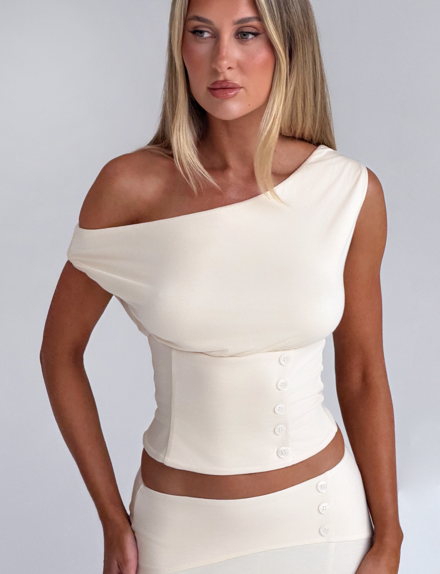 A woman with long, straight blonde hair wears the TIGERMIST ALYSSA OFF SHOULDER TOP in cream with a matching skirt featuring button details and a corseted waist, standing against a plain light background.