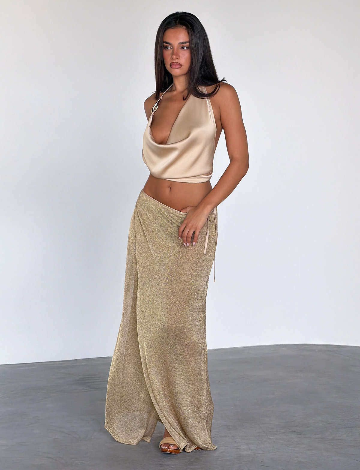 A woman with long dark hair and neutral makeup wears the TIGERMIST CYRENE SATIN COWL TOP in gold, styled with a long shimmery gold skirt and sandals. She stands indoors against plain white walls and a concrete floor.