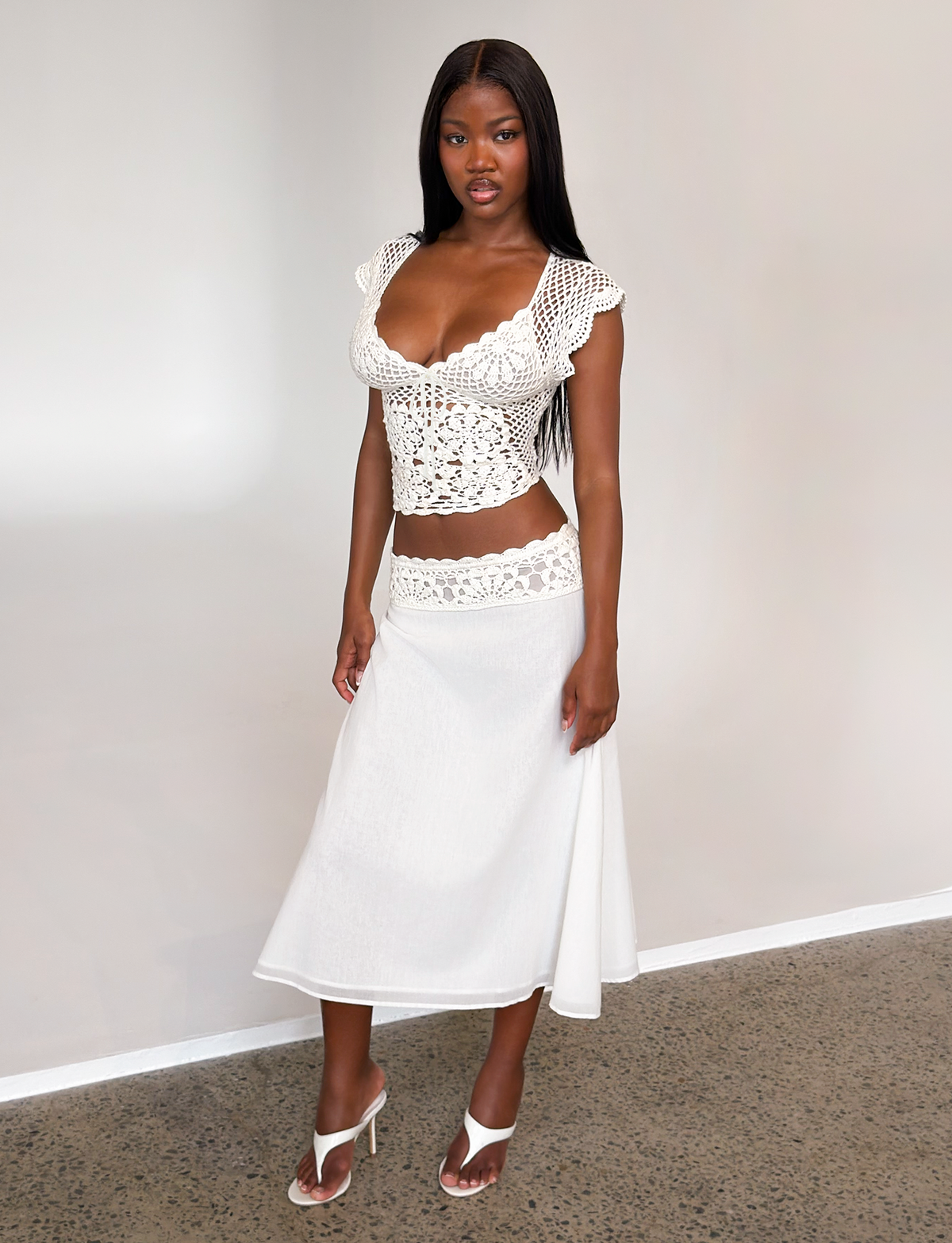 A woman stands indoors on a polished concrete floor, wearing the TIGERMIST SASHA CROCHET MIDI SKIRT in ivory, paired with a white crocheted crop top and white heeled sandals against a plain backdrop.